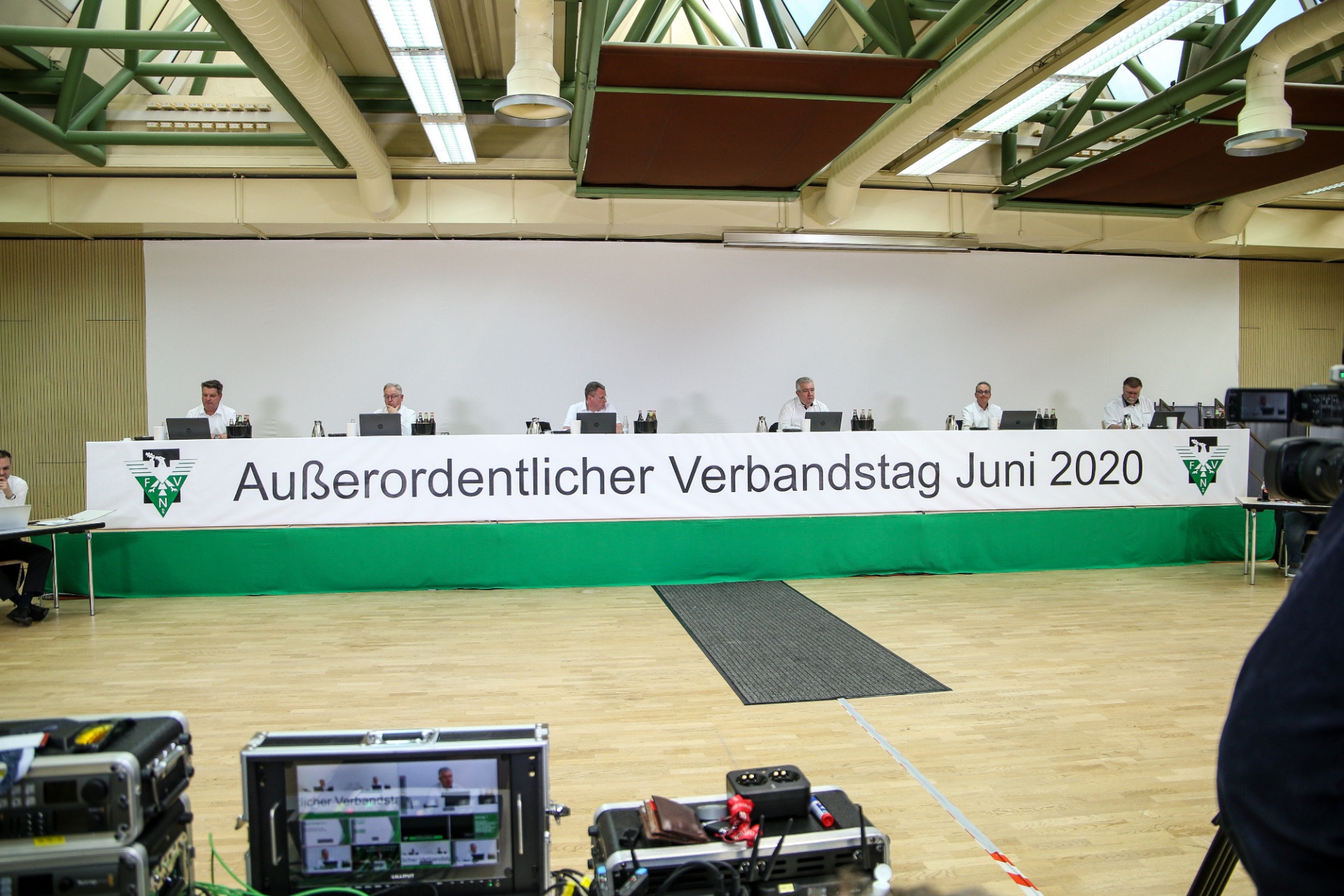 [{"clang_id":1,"value":"Reihe von Personen an einem langen Tisch mit grünem Tuch. Im Hintergrund eine Sporthalle, Schriftzug \"Außerordentlicher Verbandstag Juni 2020\"."}]