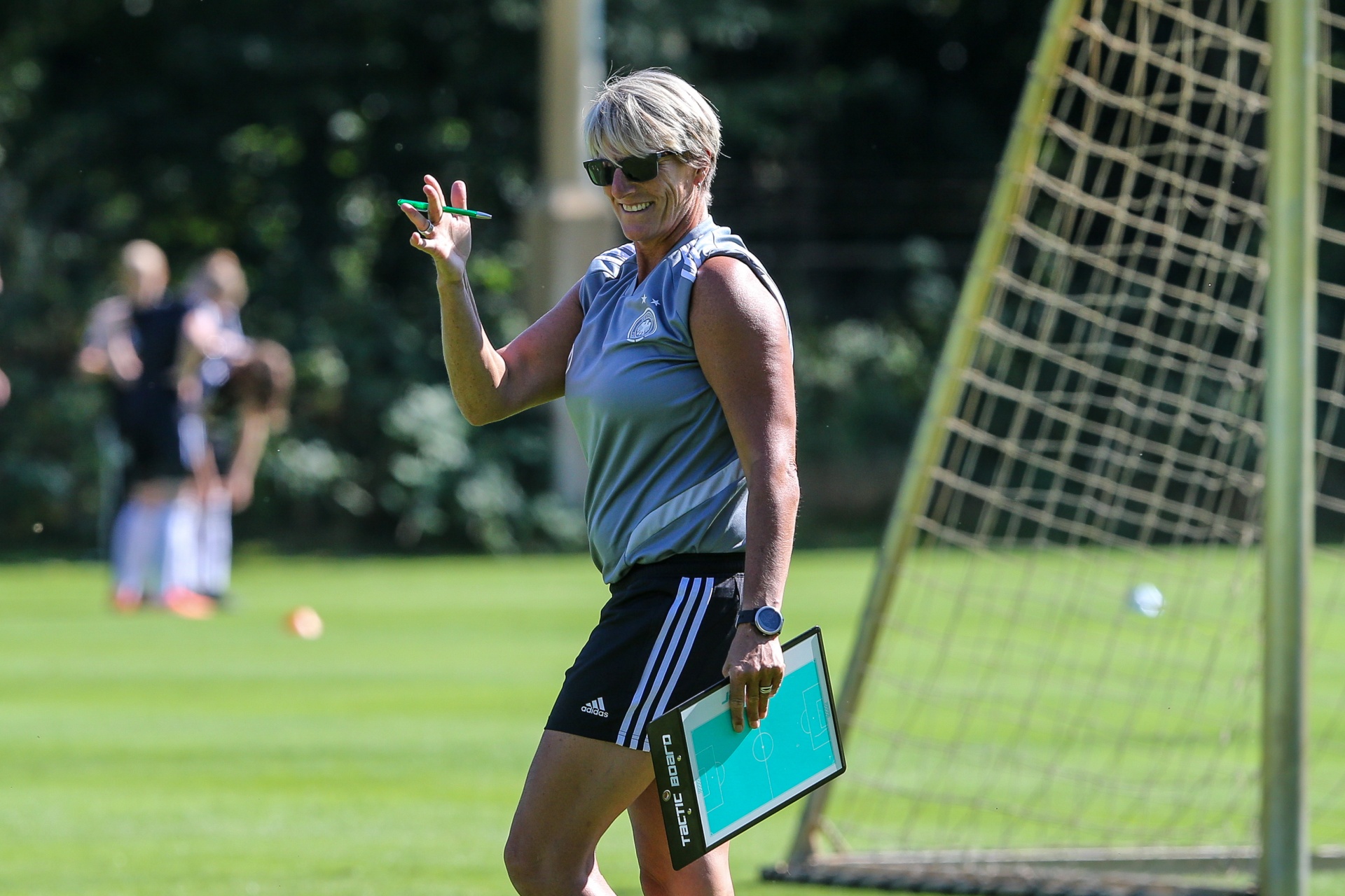 [{"clang_id":1,"value":"Frau mit Kurzhaarfrisur, Sonnenbrille und Sportkleidung steht auf einem Fußballfeld. Sie hält einen Stift und eine Taktiktafel. Im Hintergrund Spieler."}]