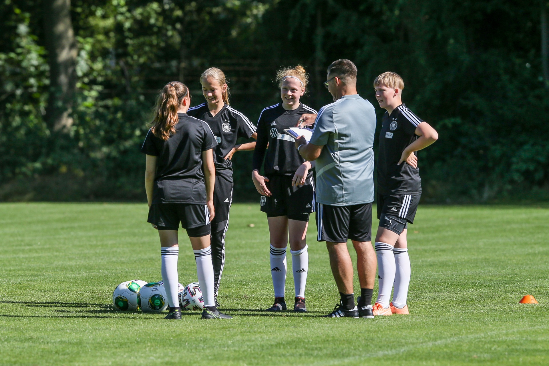 [{"clang_id":1,"value":"Fußballteam im Gespräch auf grünem Rasen. Fünf Jugendliche, überwiegend Mädchen, in Trainingsanzügen stehen um einen Trainer herum. Fußbälle in der Nähe."}]