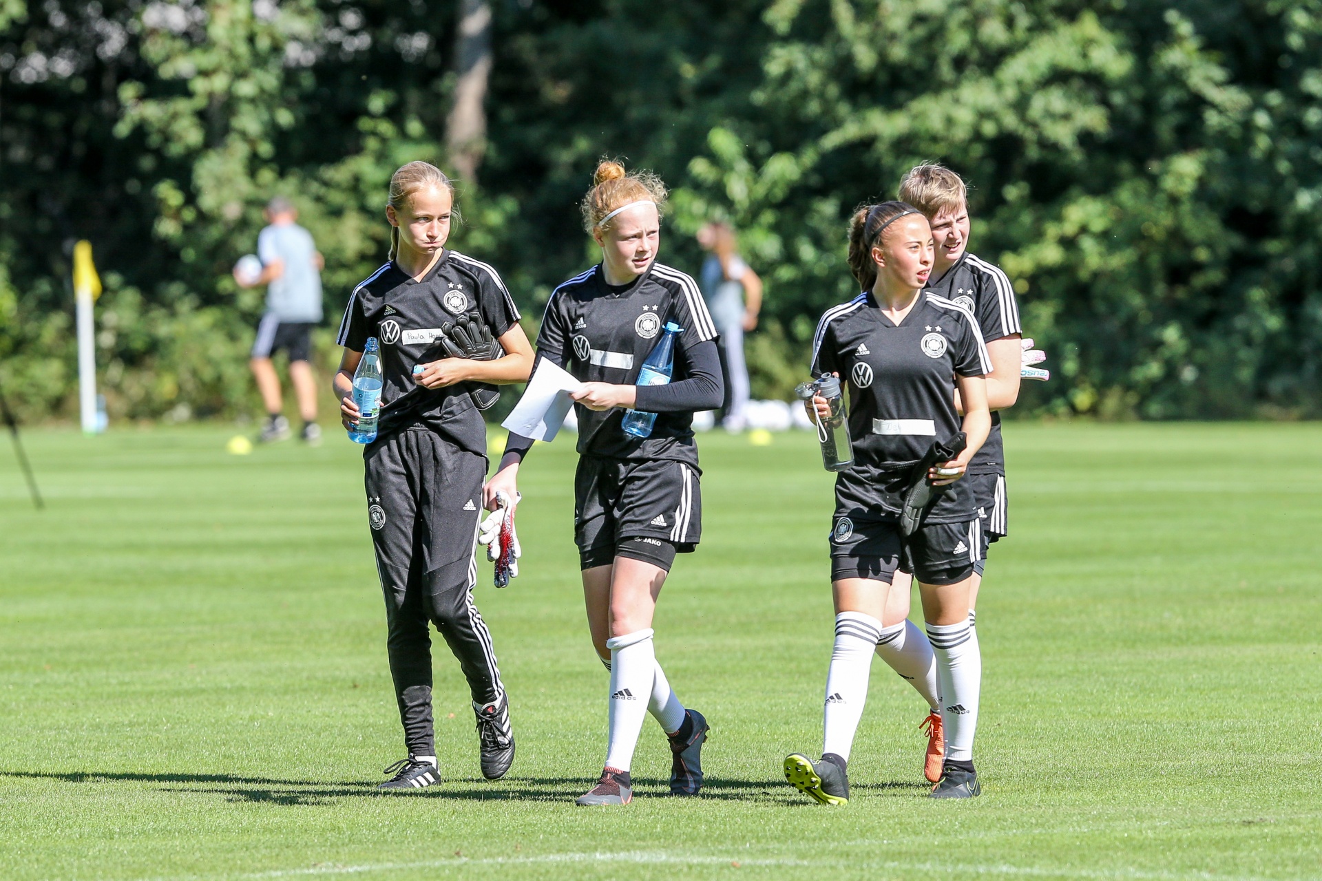 [{"clang_id":1,"value":"Vier junge Fußballspielerinnen in schwarzen Trainingsanzügen gehen auf einem grünen Rasenplatz. Jede trägt eine Wasserflasche. Im Hintergrund Bäume und andere Sportler."}]