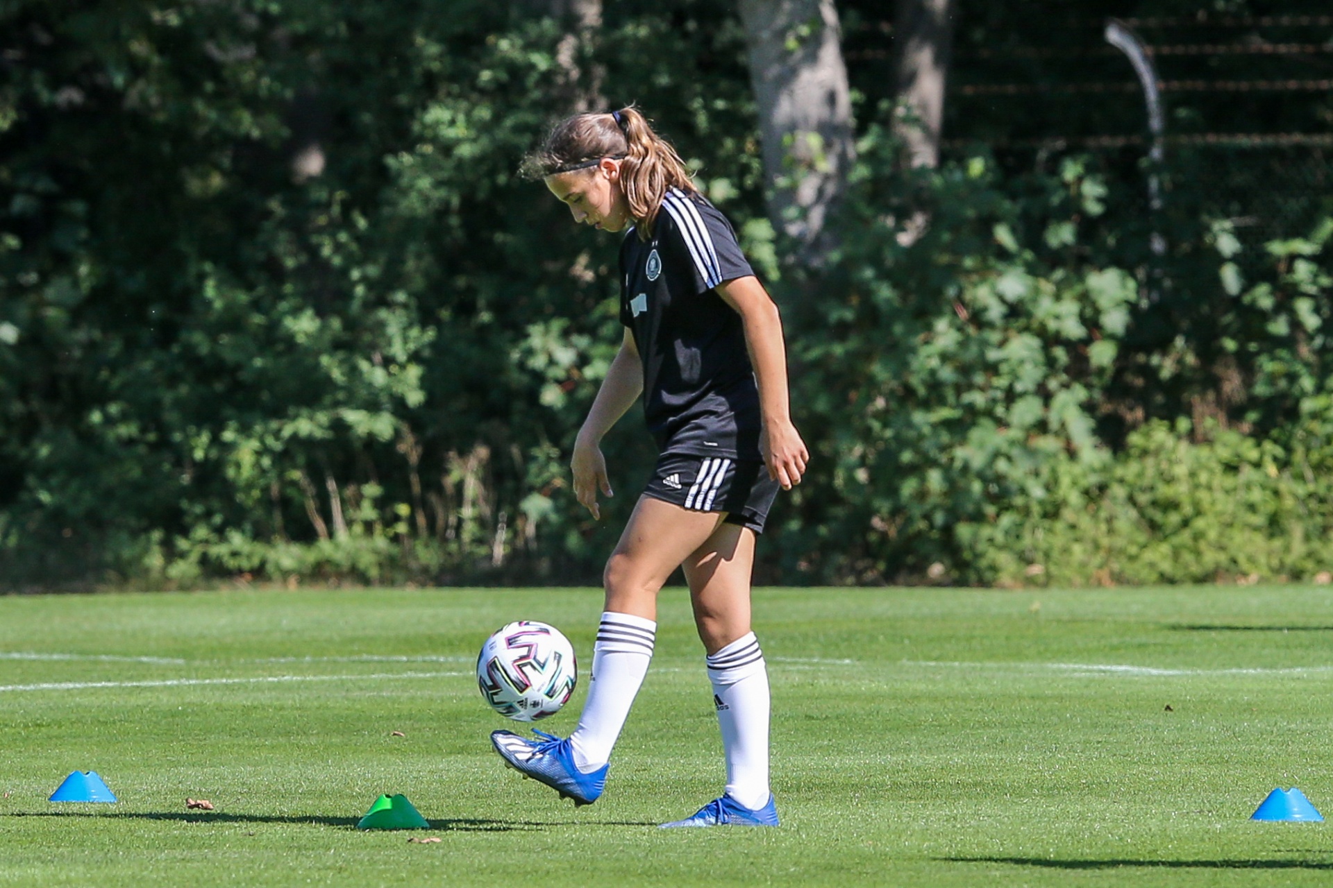 [{"clang_id":1,"value":"Fußballspielerin mit dunklem Pferdeschwanz dribbelt auf grünem Rasen einen Fußball. Im Hintergrund Bäume und blaue Markierungshütchen."}]