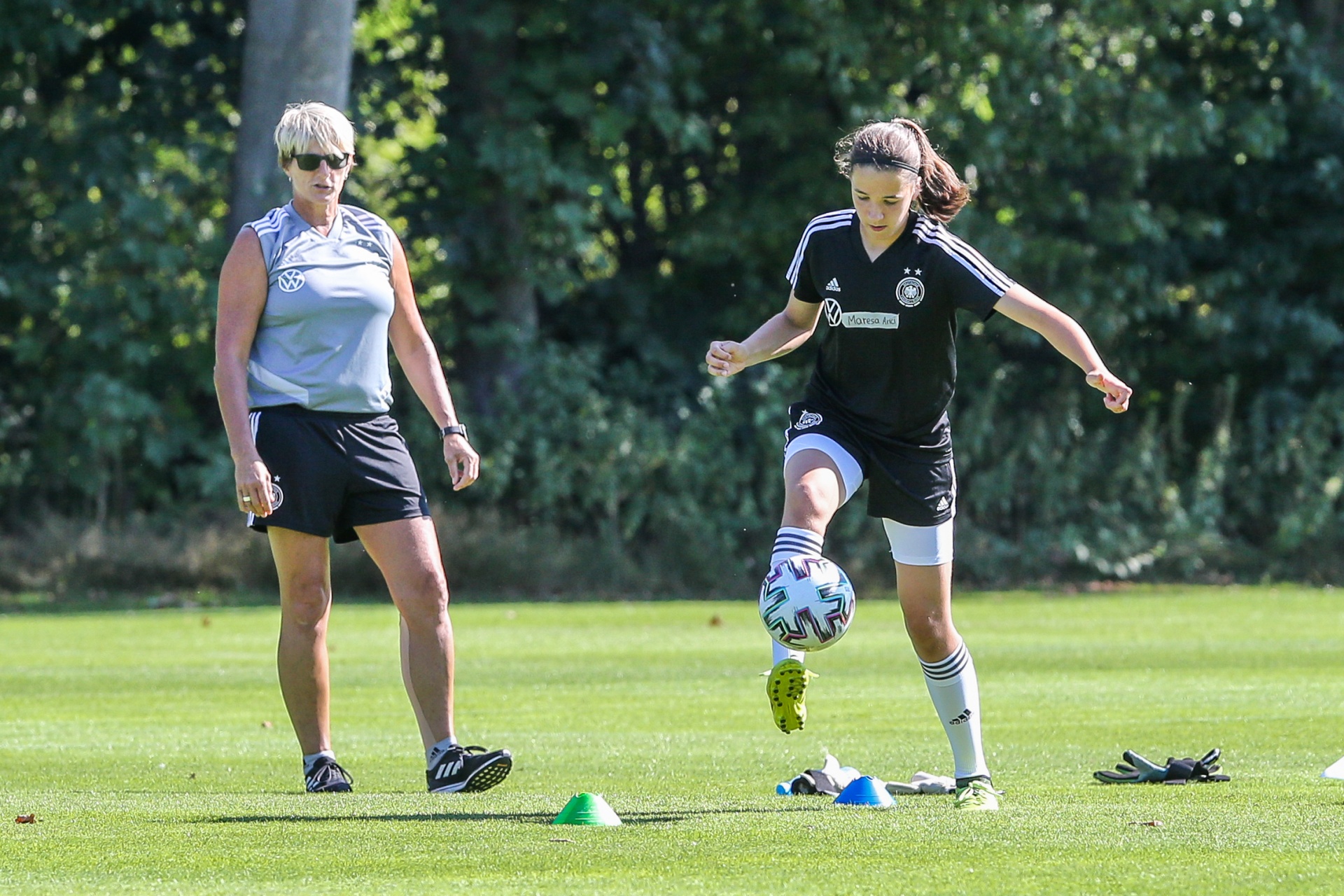 [{"clang_id":1,"value":"Fußballtrainerin steht am Spielfeldrand, während eine junge Spielerin den Ball jongliert. Grüne Wiese und Bäume im Hintergrund."}]