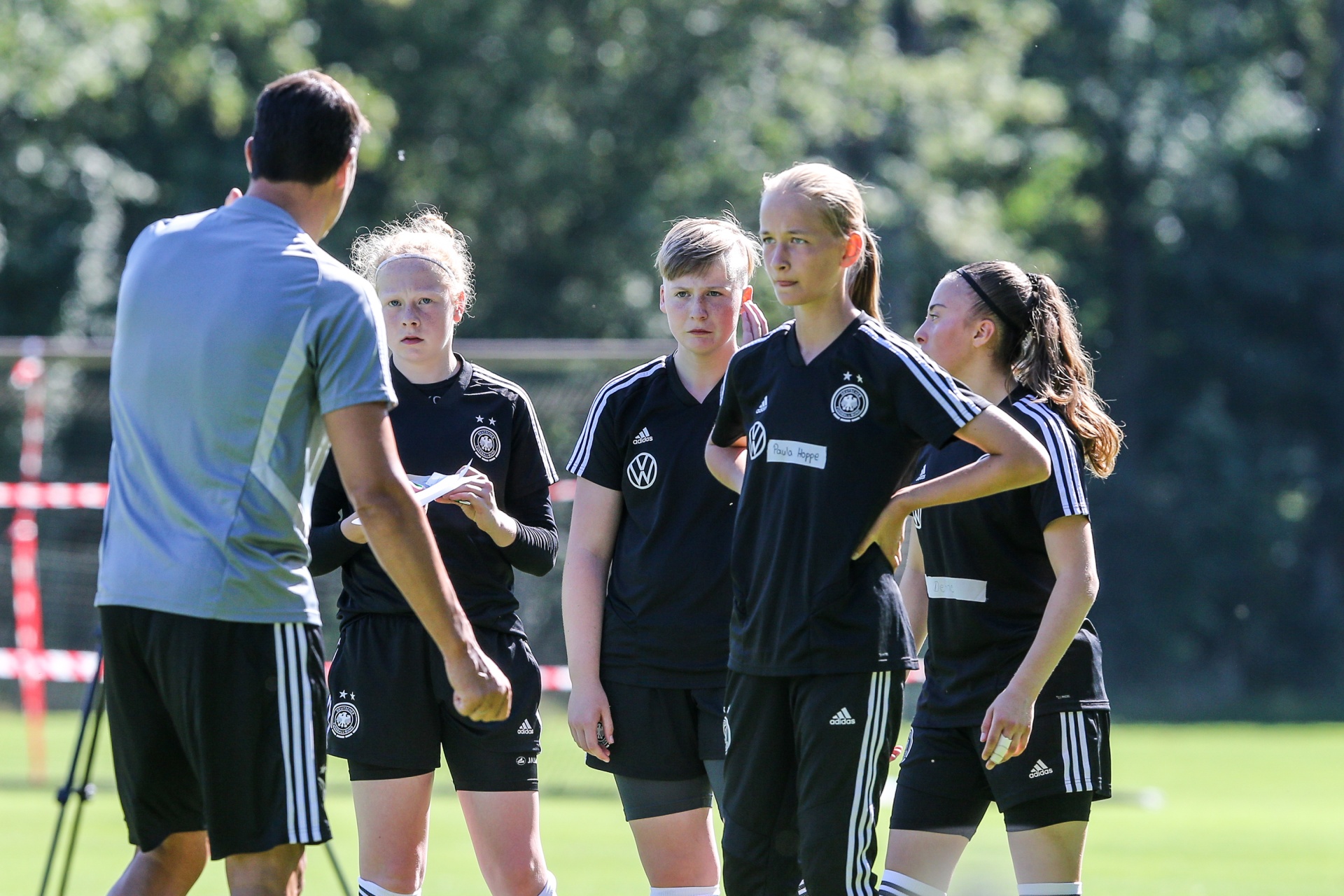 [{"clang_id":1,"value":"Fußballtrainer spricht vier jugendliche Spielerinnen in Trainingskleidung auf einem Rasenplatz an. Im Hintergrund Bäume und Fußballtor."}]