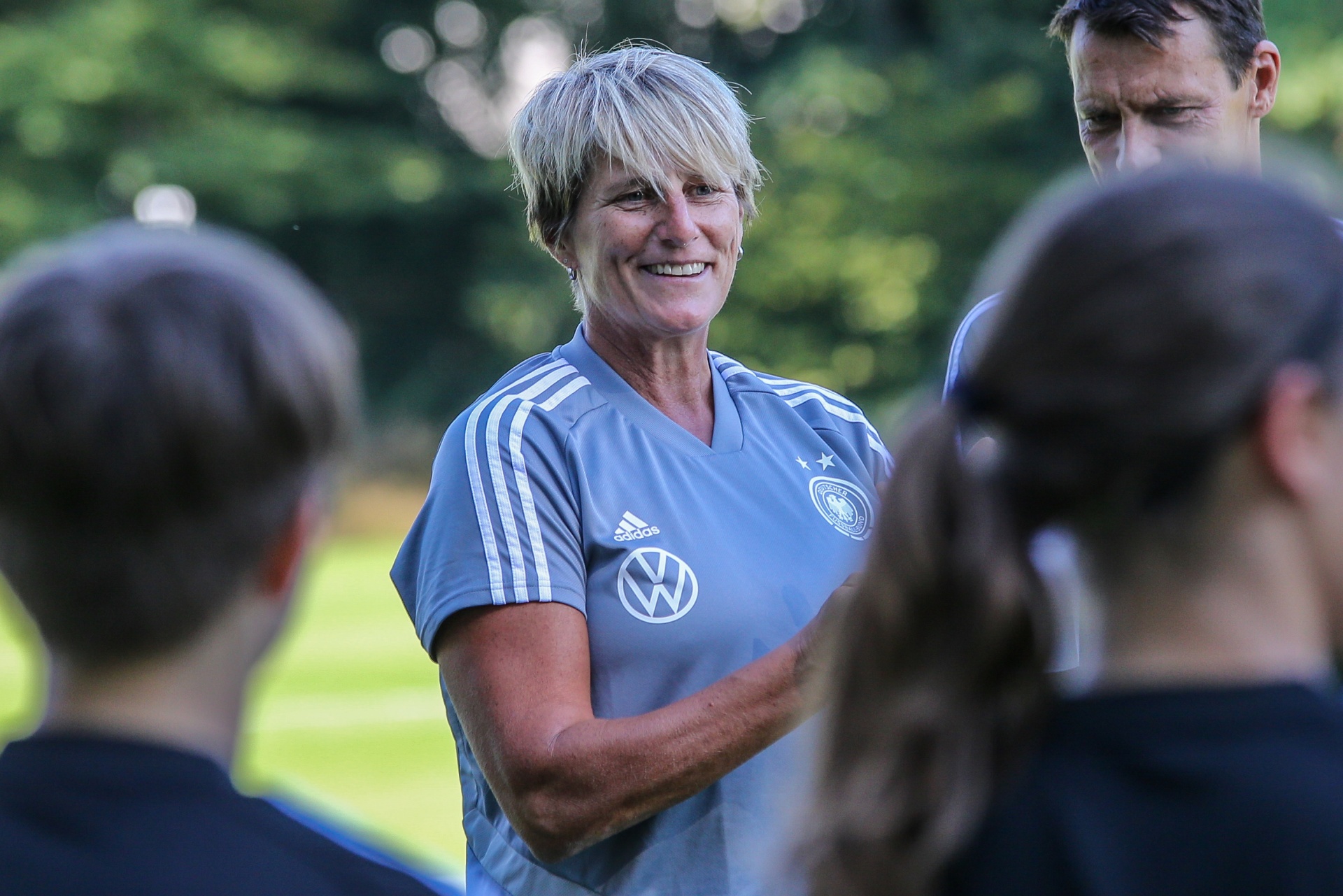 [{"clang_id":1,"value":"Frau mit kurz geschnittenem Haar lächelt in einem blauen Trainingsanzug, umgeben von Kindern auf einem Fußballfeld. Im Hintergrund ein weiterer Trainer."}]