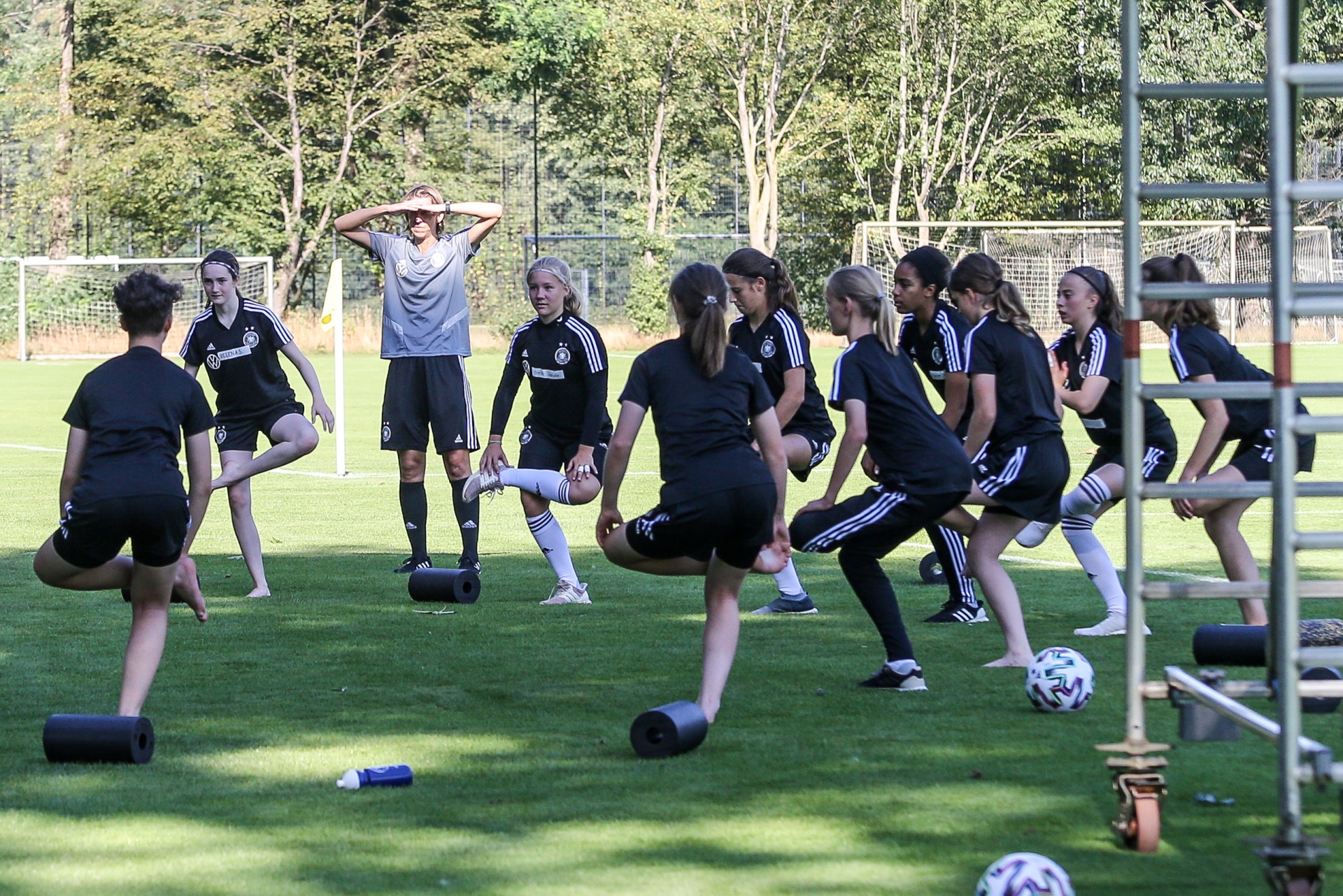 [{"clang_id":1,"value":"Fußballspielerinnen dehnen auf grünem Rasen. Eine Trainerin steht inmitten der Gruppe. Im Hintergrund ein Zaun und Bäume."}]