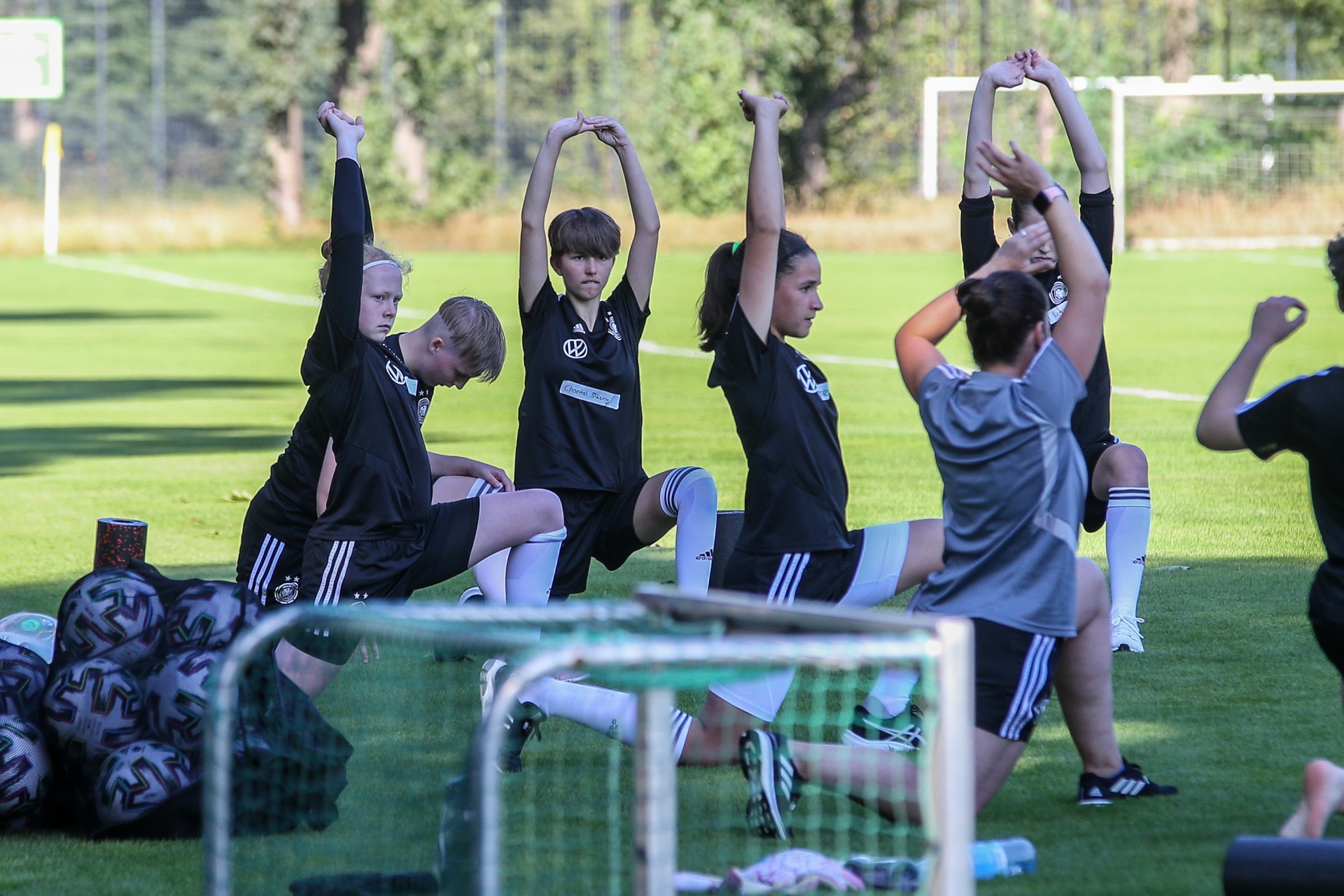[{"clang_id":1,"value":"Fußballspieler*innen im Trainingsanzug dehnen sich auf einem Fußballfeld. Einige knien, andere stehen in der Ausfallposition mit erhobenen Armen. Im Vordergrund Fußballtore und -bälle."}]
