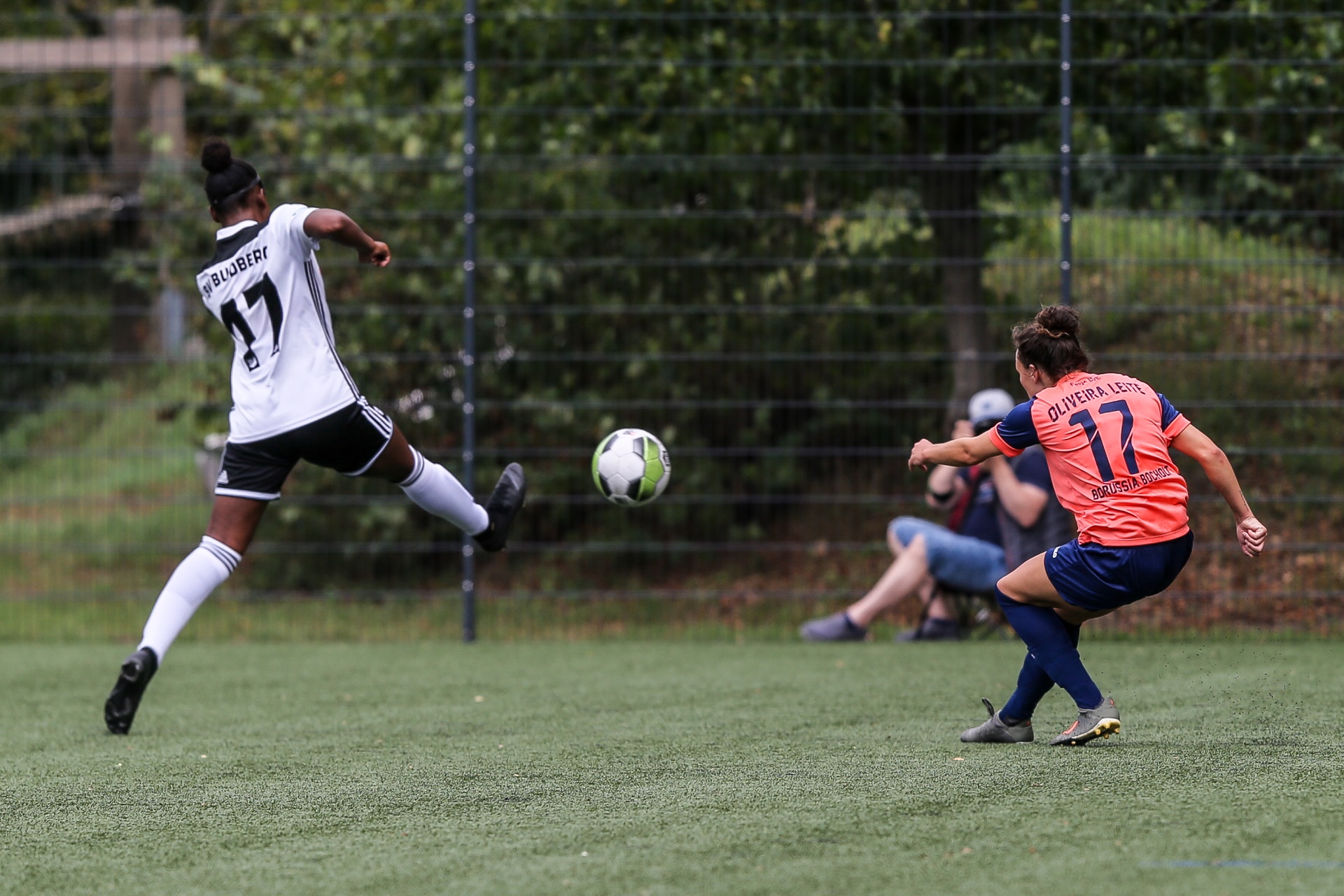 [{"clang_id":1,"value":"Zwei Fußballspielerinnen auf grünem Rasen. Links eine Spielerin mit Nummer 17 in schwarz-weißem Trikot beim Schießen, rechts eine Spielerin mit rotem Trikot und dem Namen \"Oliveira Leite\". Im Hintergrund weitere Personen."}]