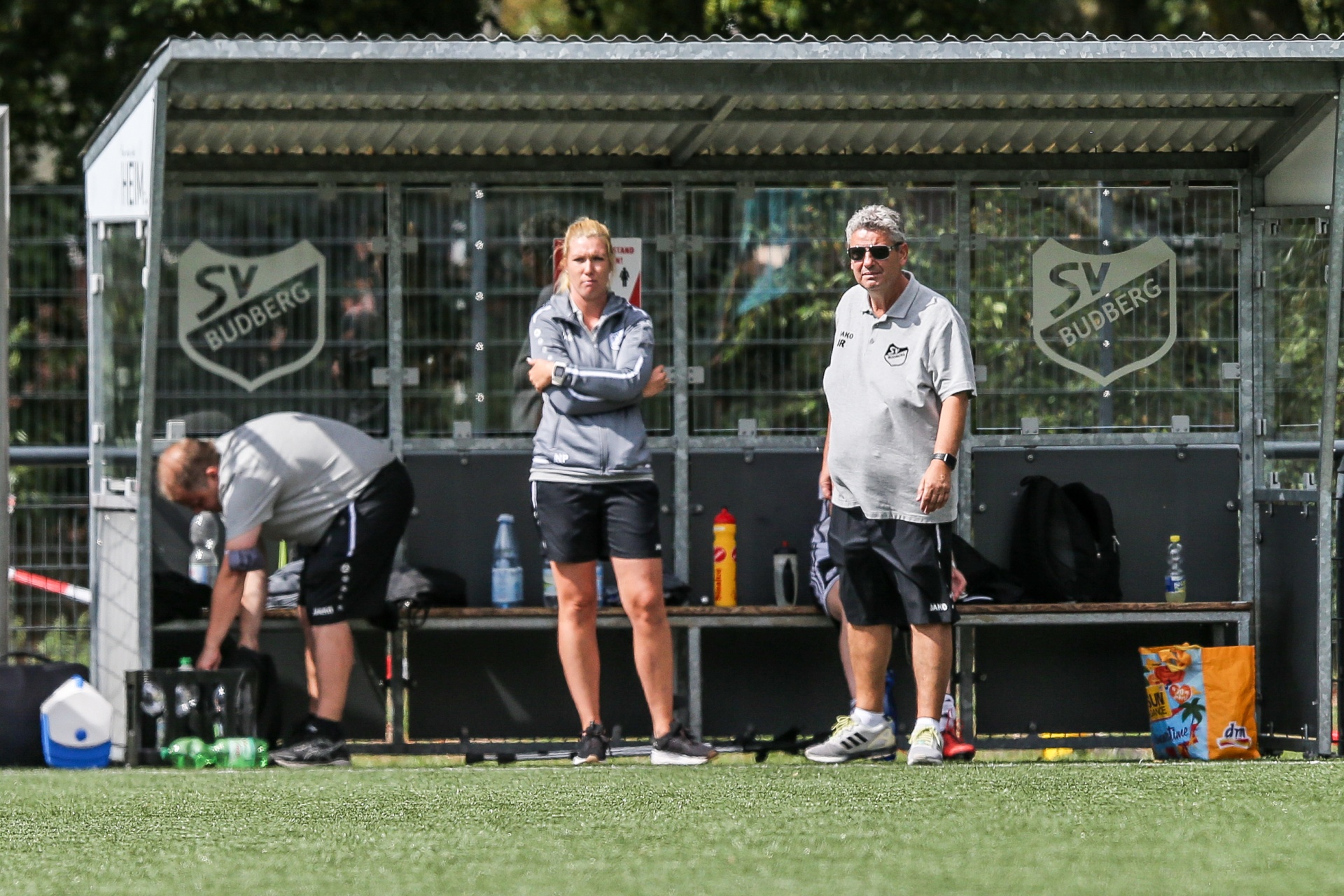 [{"clang_id":1,"value":"Fußballtrainer und -betreuer vor einer Trainerbank mit Vereinslogo \"SV Budberg\". Im Hintergrund grünes Fußballfeld und Bäume."}]