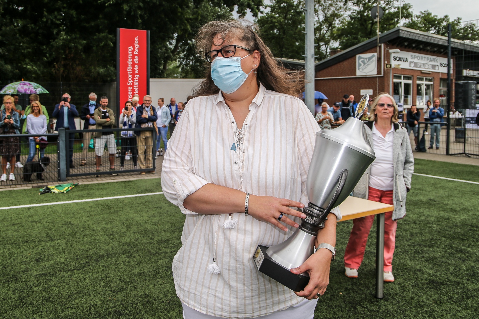 [{"clang_id":1,"value":"Frau mit Maske hält Trophäe im Sportpark. Im Hintergrund Zuschauer und Gebäude. Rasenplatz im Vordergrund."}]