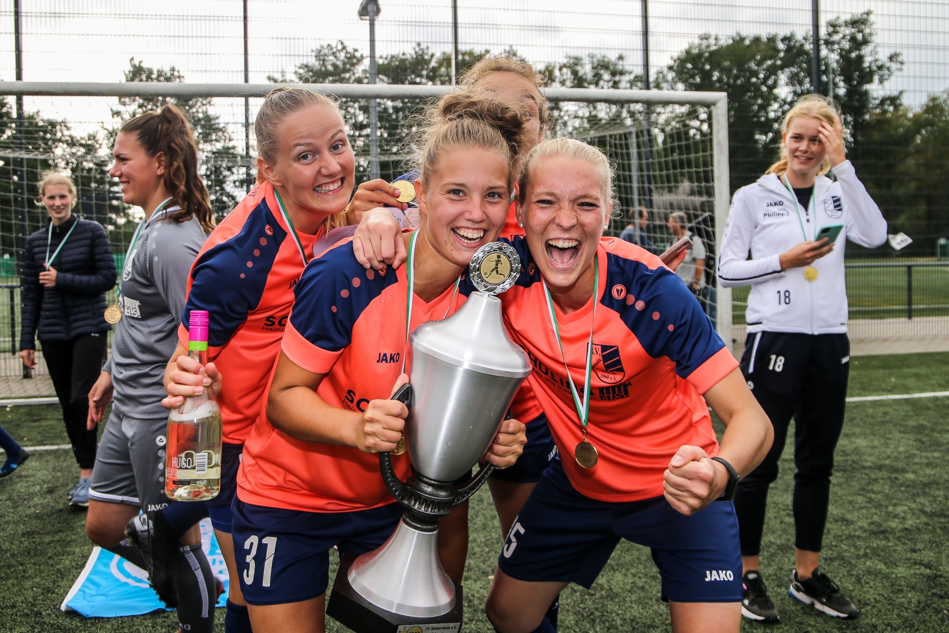 [{"clang_id":1,"value":"Fußballspielerinnen in orange-blauen Trikots feiern mit Pokal und Medaillen. Im Hintergrund steht ein Fußballtor und weitere Teammitglieder."}]