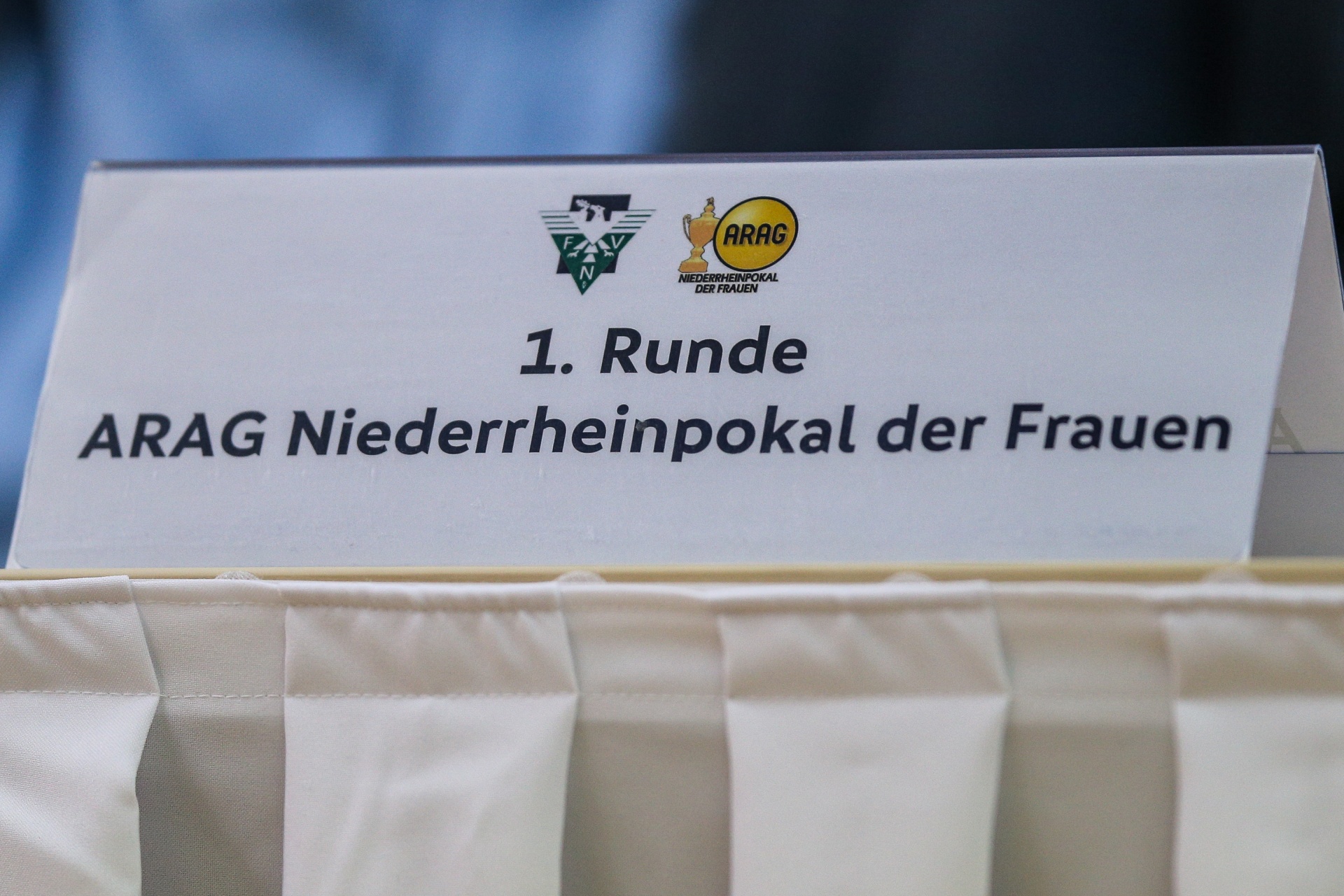 [{"clang_id":1,"value":"Weißer Karton mit Aufdruck \"1. Runde ARAG Niederrheinpokal der Frauen\". Darauf Logos von FVN und ARAG. Darunter sind weiße Gegenstände zu sehen."}]
