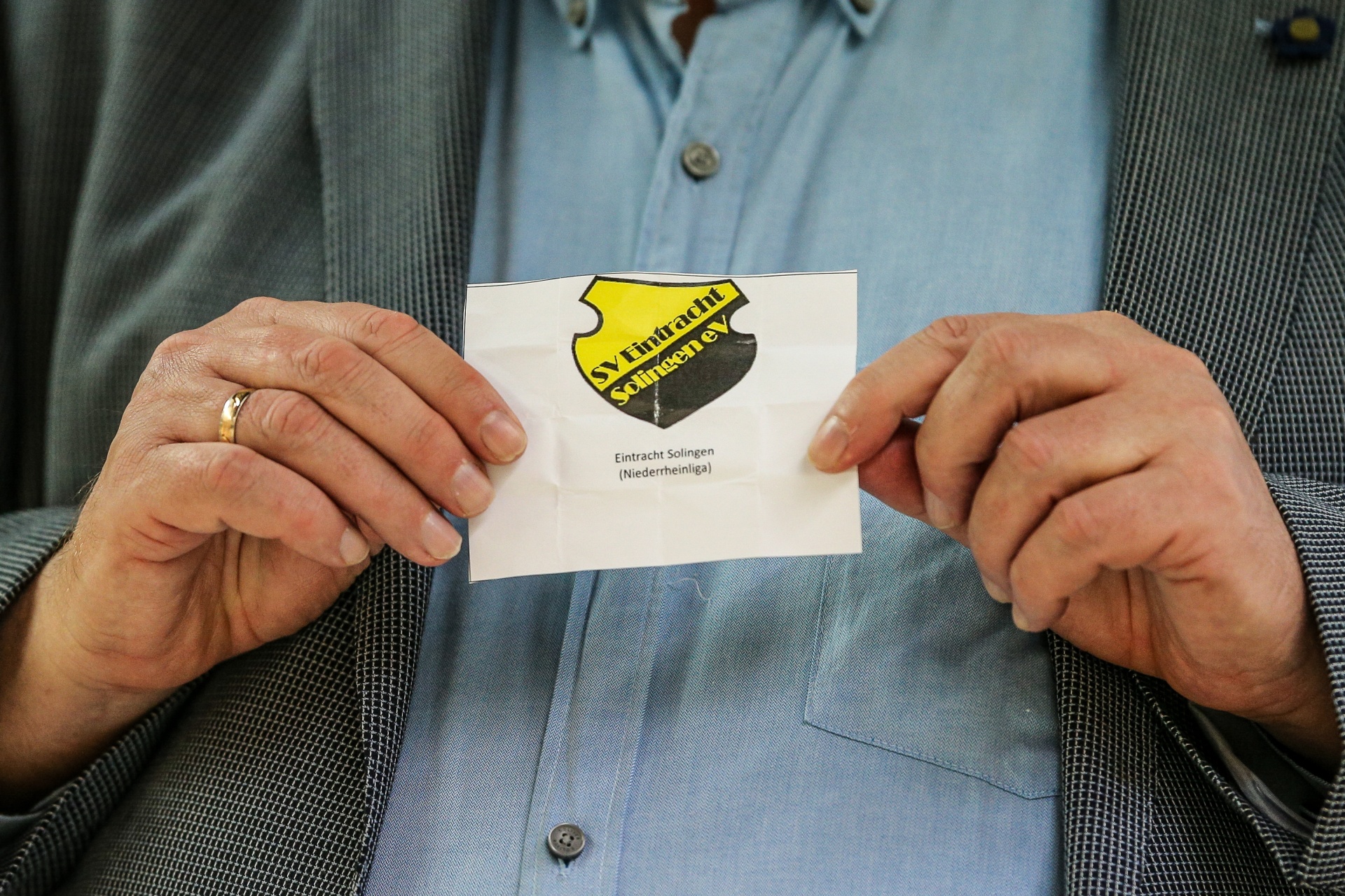 [{"clang_id":1,"value":"Ältere Hände halten eine weiße Karte mit dem Logo des SV Eintracht Solingen e.V. vor einem Mann mit hellbrauner Jacke."}]