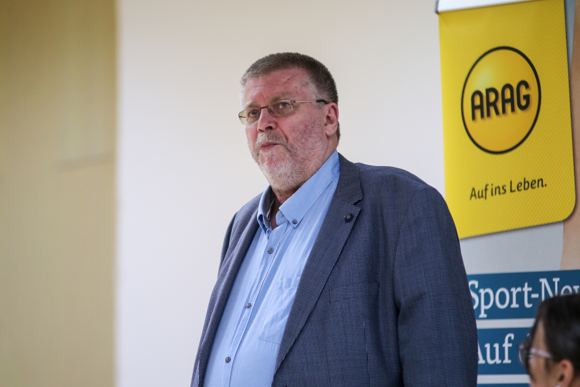 [{"clang_id":1,"value":"Mann mit Bart und Brille im Anzug vor gelbem Banner mit Schriftzug \"ARAG\"."}]