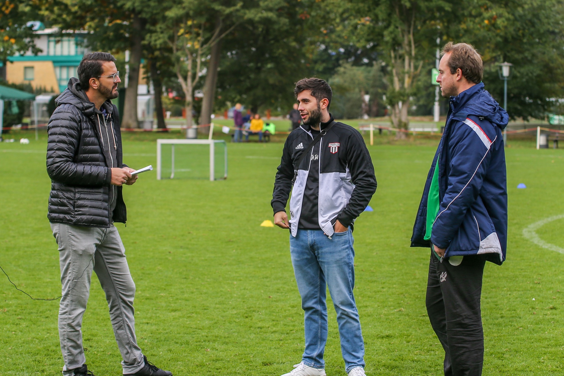 [{"clang_id":1,"value":"Drei Männer stehen auf einem Fußballfeld und unterhalten sich. Links trägt einer eine dunkle Jacke, in der Mitte ein sportlicher Pullover und rechts eine blaue Trainingsjacke. Im Hintergrund ein Fußballtor und Bäume."}]