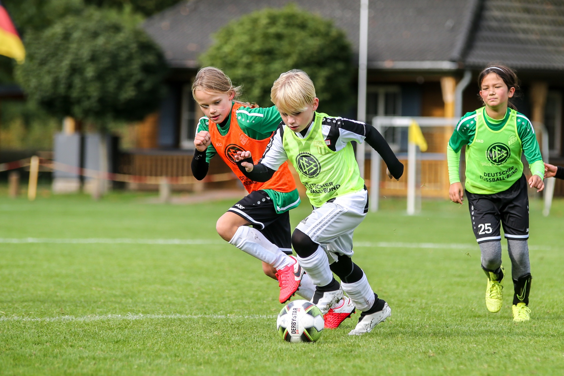 [{"clang_id":1,"value":"Drei Kinder spielen Fußball auf einem grünen Rasenplatz. Ein Junge dribbelt den Ball, ein Mädchen in orangefarbener Weste rennt links, ein weiteres Mädchen in grün-weißer Kleidung rechts. Im Hintergrund ein Holzgebäude."}]