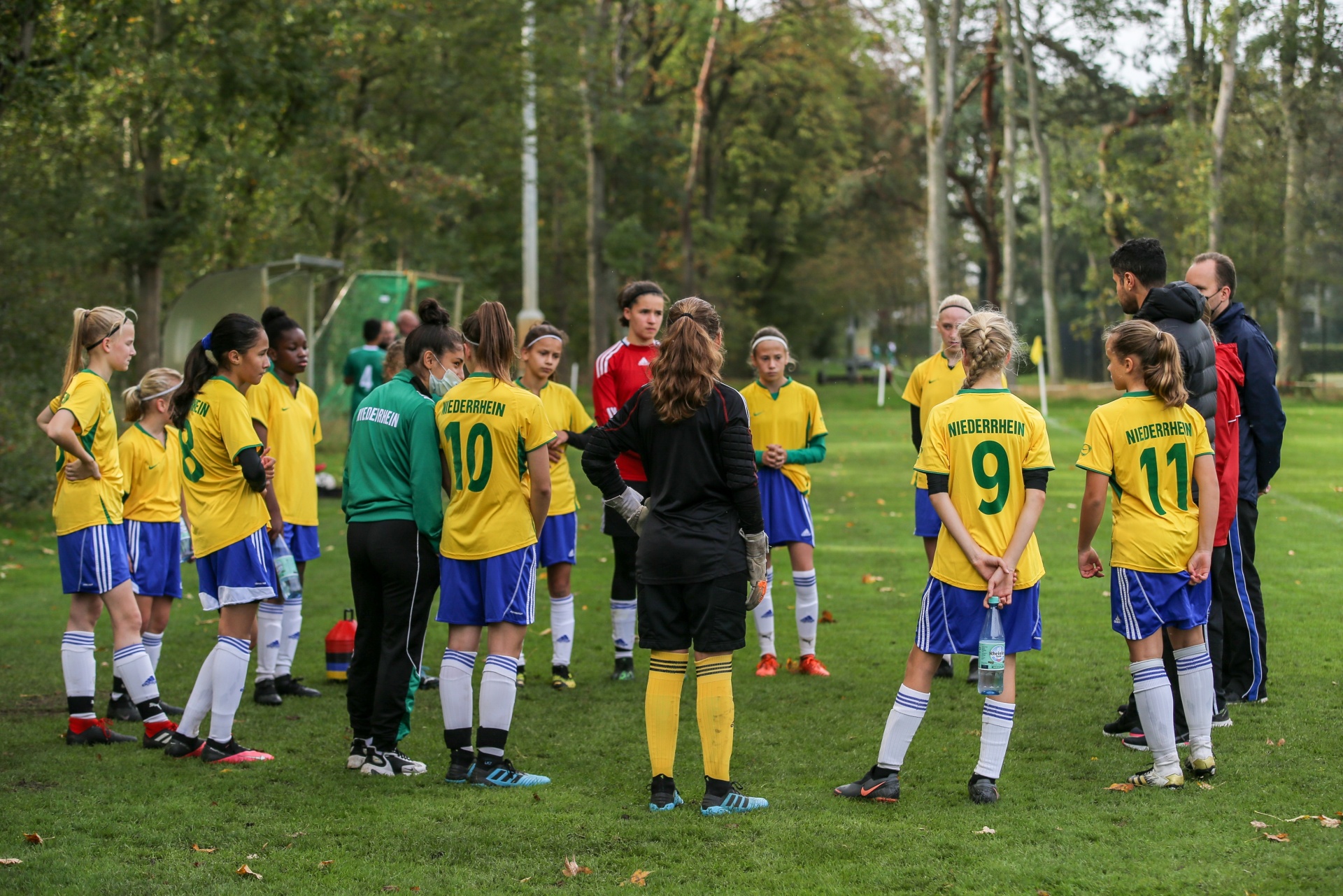 [{"clang_id":1,"value":"Fußballteam im Kreis. Spielerinnen in gelben und grünen Trikots stehen um eine Trainerin auf einem Rasenplatz. Bäume im Hintergrund."}]