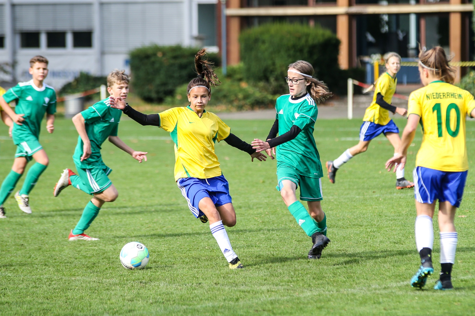 [{"clang_id":1,"value":"Fußballspiel: Jugendmannschaften in grünen und gelben Trikots kämpfen um den Ball auf dem Rasenplatz. Mehrere Spieler in Bewegung. Gebäude im Hintergrund."}]