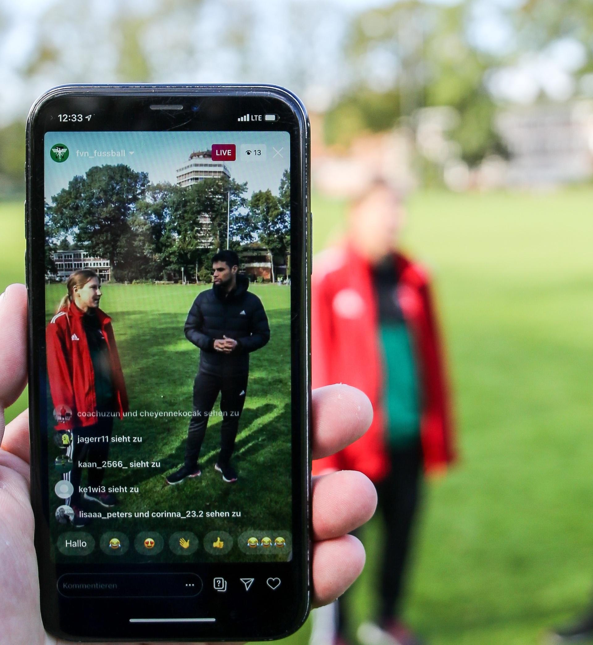 [{"clang_id":1,"value":"Smartphone zeigt Live-Übertragung eines Fußballtrainings. Zwei Männer in Trainingsanzügen stehen auf dem Rasen, umgeben von Zuschauern. Chat-Nachrichten sind sichtbar."}]