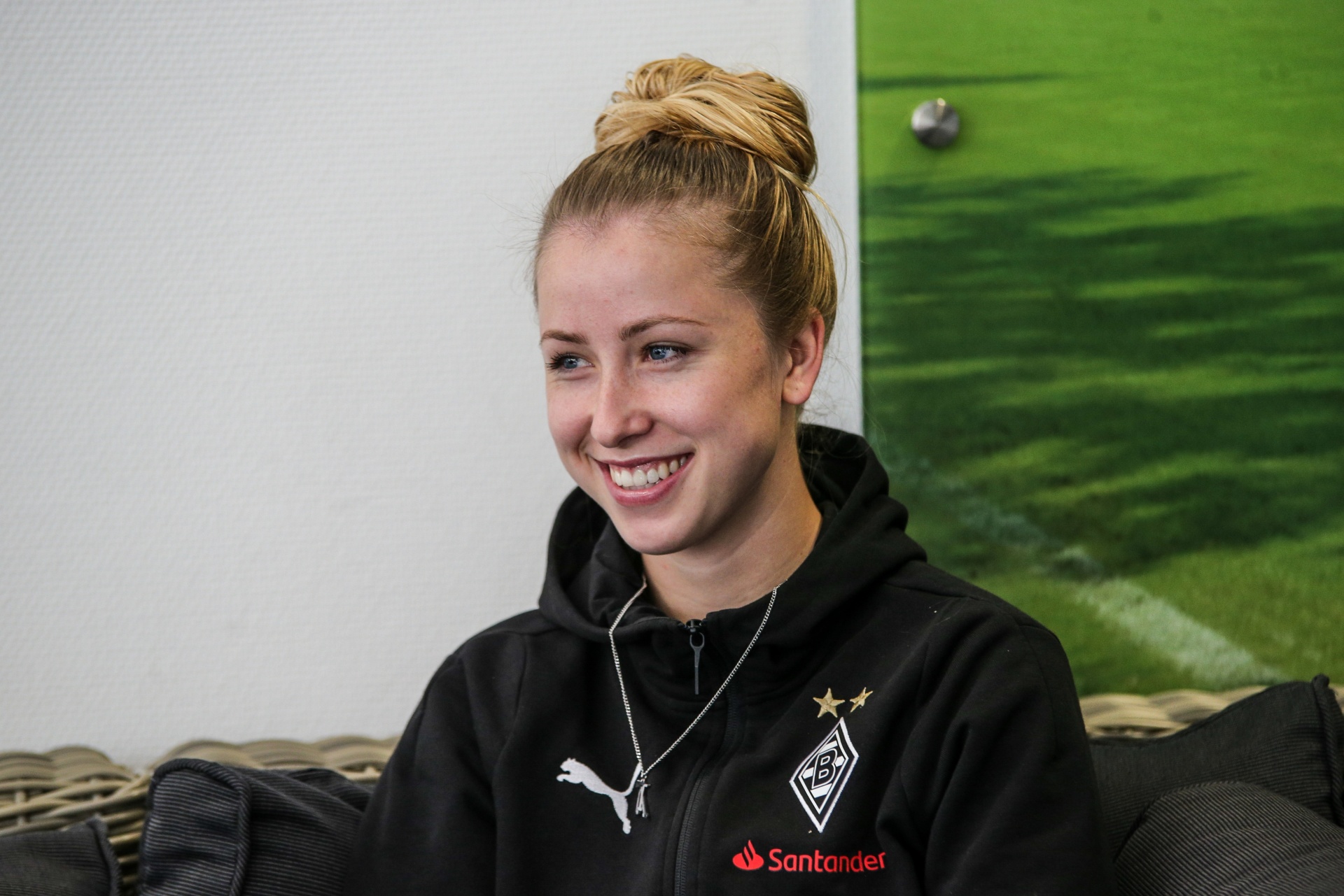 [{"clang_id":1,"value":"Lächelnde blonde Frau mit hochgestecktem Haar in schwarzer Trainingsjacke mit Borussia Mönchengladbach Logo und Santander Schriftzug. Hintergrund: heller Raum, grüner Teppich."}]