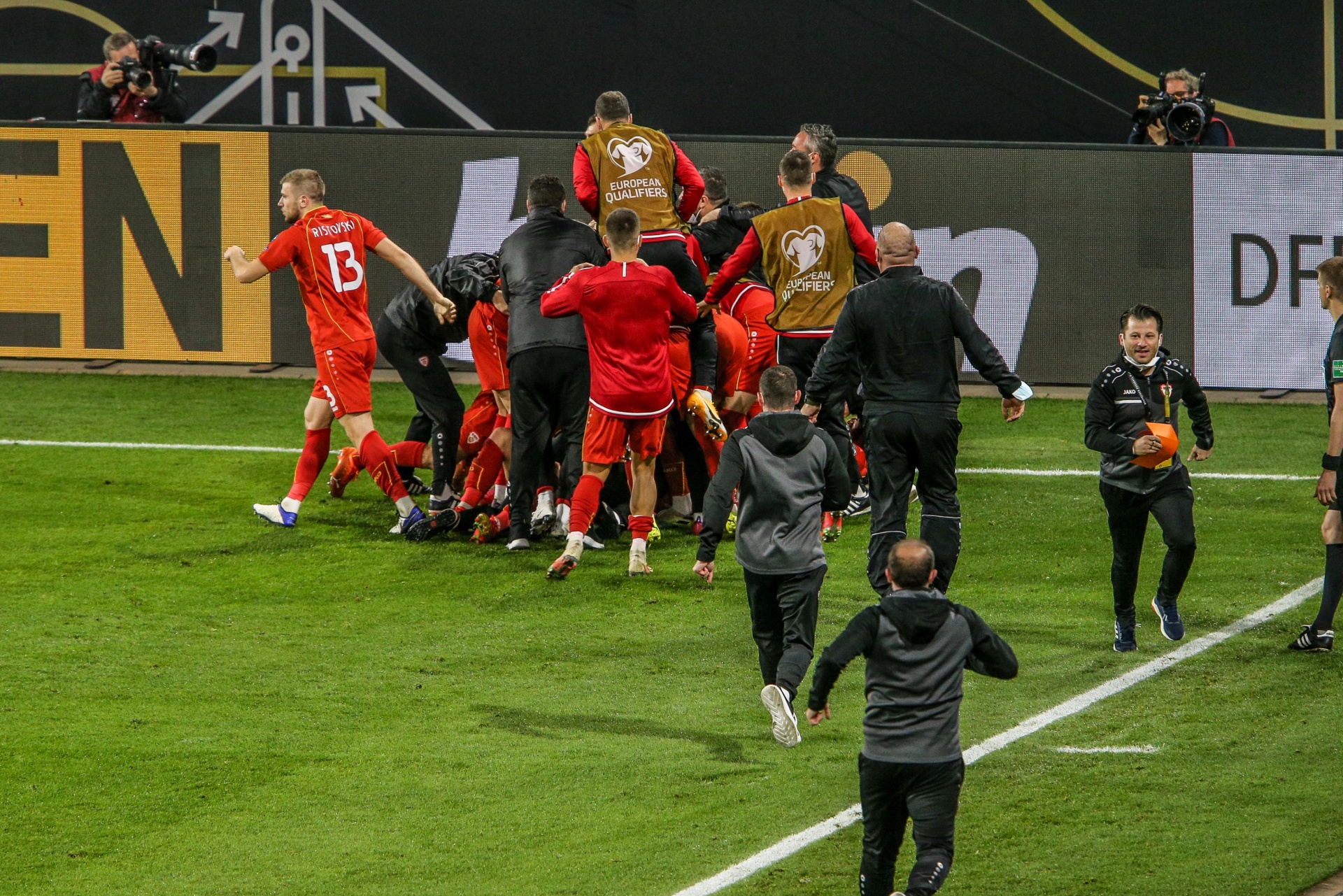 [{"clang_id":1,"value":"Fußballspieler in roten Trikots feiern auf dem Rasen, umgeben von Ordnern und weiteren Personen. Im Hintergrund eine Anzeigetafel mit \"EN\" und \"DR\"."}]