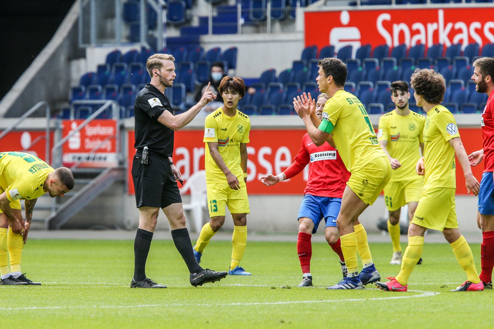 [{"clang_id":1,"value":"Fußballspiel: Schiedsrichter mit erhobener roter Karte vor Spielern in gelben und roten Trikots. Stadion im Hintergrund."}]