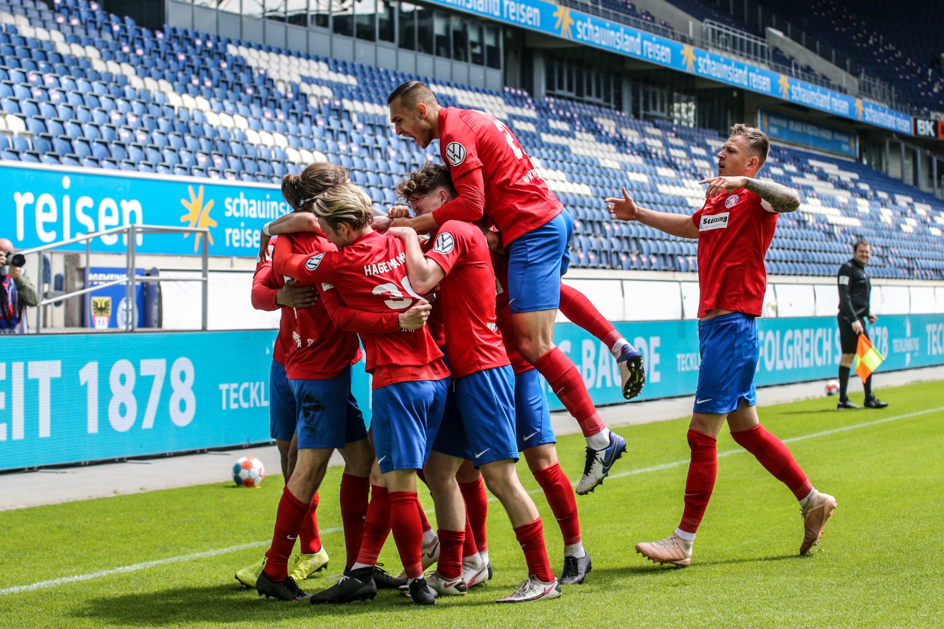 [{"clang_id":1,"value":"Fußballspieler feiern Tor. Gruppe in rot-blauen Trikots jubelt auf grünem Rasen vor Stadiontribüne. Ein Spieler wird hochgehoben."}]