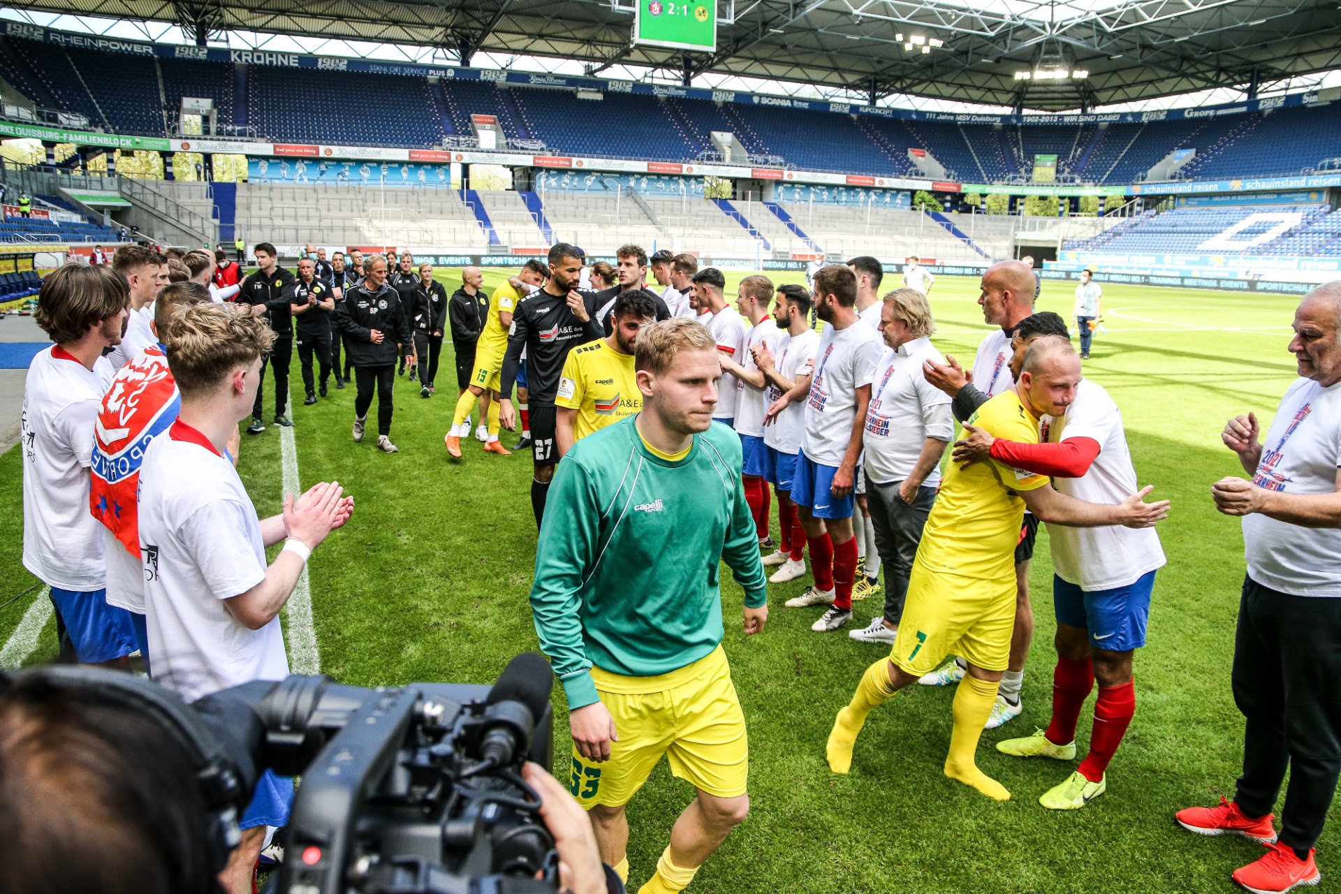 [{"clang_id":1,"value":"Fußballstadion: Spieler in Vereinskleidung stehen im Kreis, applaudieren und begrüßen sich. Einige tragen Trainingsanzüge. Im Hintergrund leere Zuschauerränge."}]