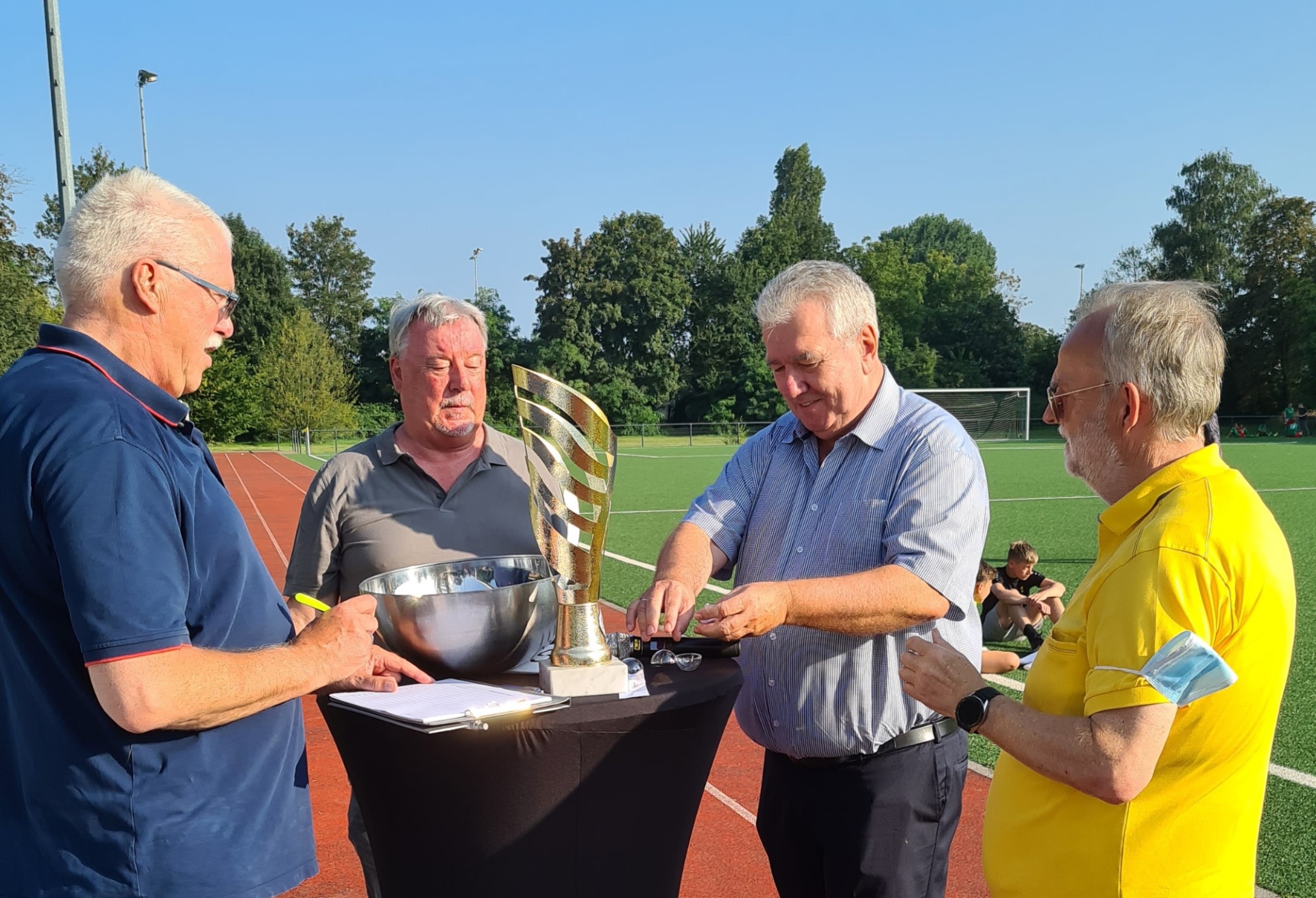 [{"clang_id":1,"value":"Vier ältere Männer stehen um einen Tisch mit Pokal und Urkunde auf einem Sportplatz. Im Hintergrund ein Fußballfeld und Bäume bei strahlendem Himmel."}]
