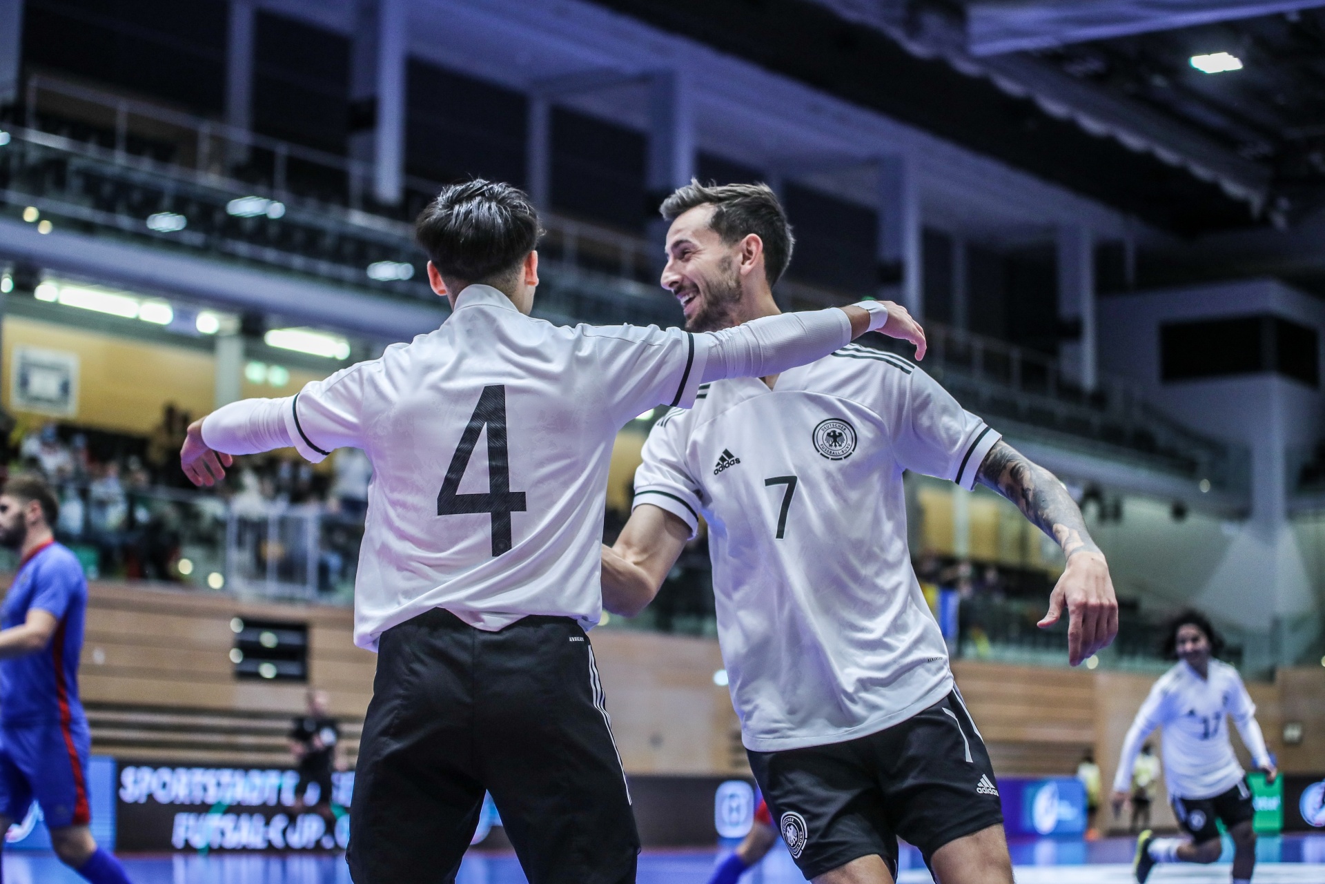 [{"clang_id":1,"value":"Zwei Futsalspieler mit Trikotnummer 4 und 7 in weißen Trikots und schwarzen Shorts feiern mit verschränkten Armen. Im Hintergrund verschwommene Zuschauer und Sportstätte."}]
