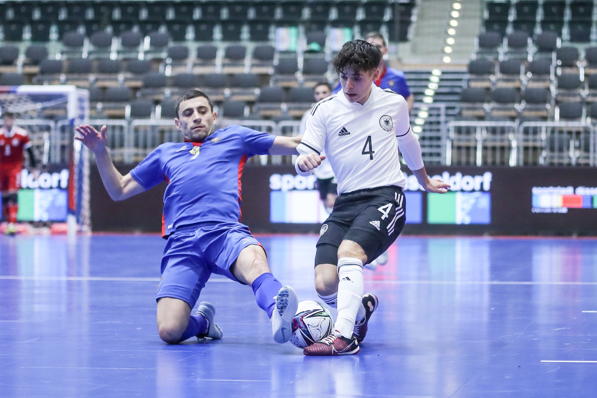 [{"clang_id":1,"value":"Futsalspiel: Spieler mit Nummer 4 in weiß-schwarzem Trikot schießt auf den Ball, während Gegenspieler in blau kniend grätscht. Hintergrund: leere Zuschauertribüne."}]
