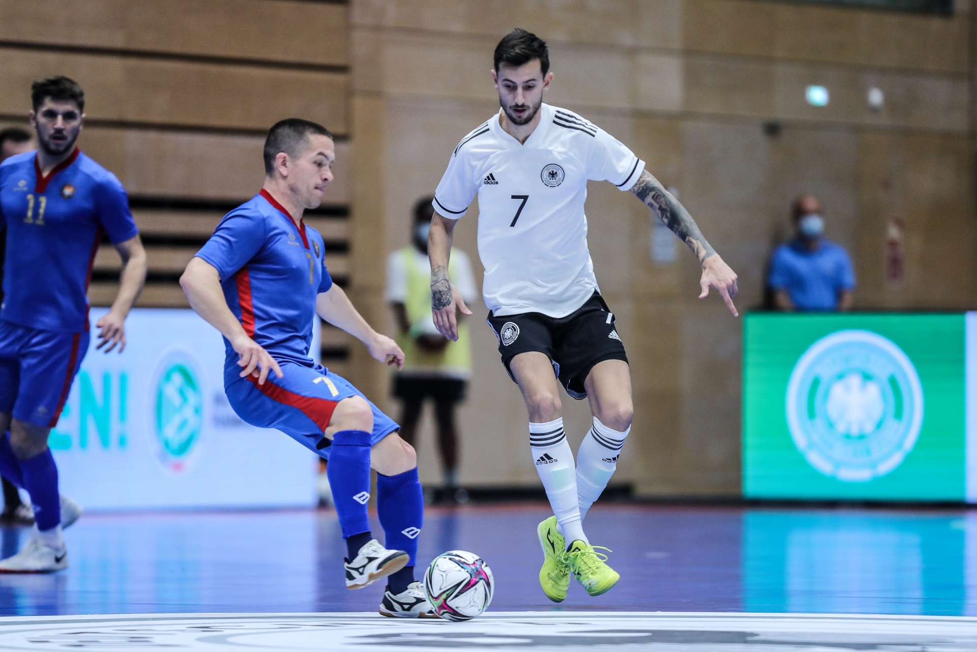 [{"clang_id":1,"value":"Futsalspiel: Spieler mit Nummer 7 im weißen Trikot dribbelt den Ball. Zwei Gegenspieler in blau-roten Trikots stehen in der Nähe. Im Hintergrund Zuschauer."}]