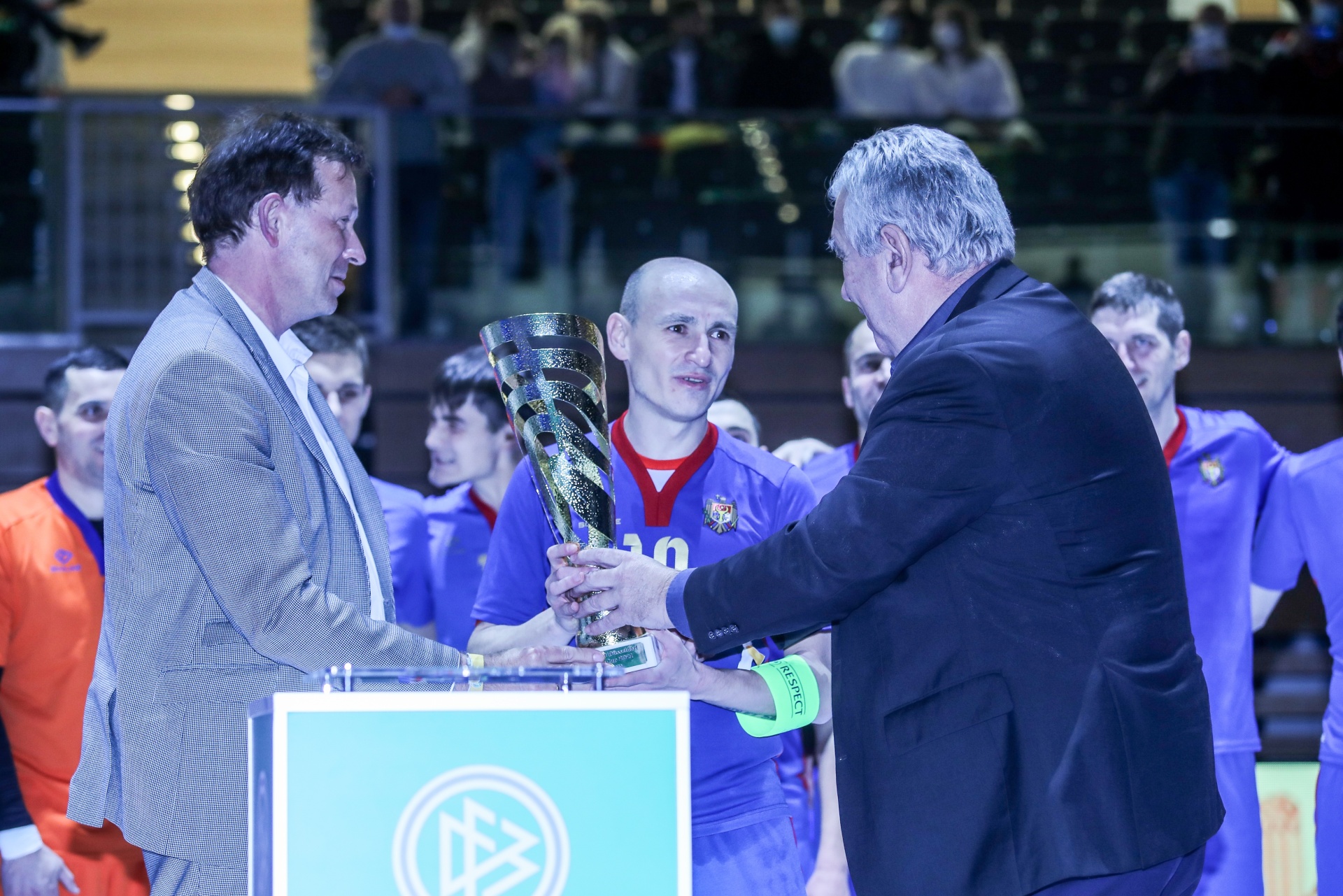 [{"clang_id":1,"value":"Zwei Männer in Anzug überreichen Pokal an Futsalspieler mit Trikotnummer 10. Im Hintergrund weitere Sportler und Zuschauer."}]