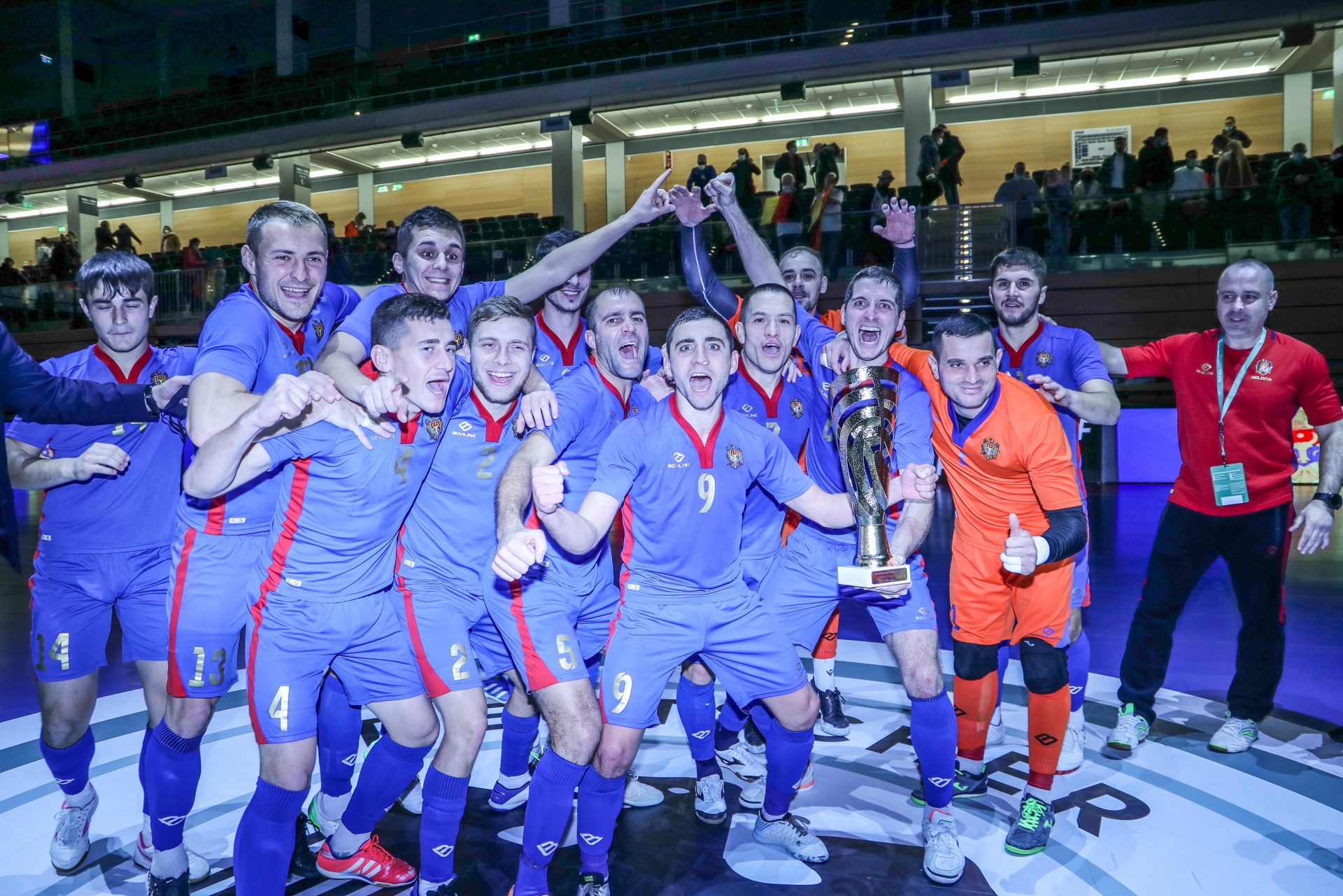 [{"clang_id":1,"value":"Mannschaft in blau-roten Trikots jubelt mit Pokal in einer Sporthalle. Im Hintergrund Zuschauer und weitere Vereinsmitglieder. Ein Spieler trägt orangefarbenes Trikot."}]