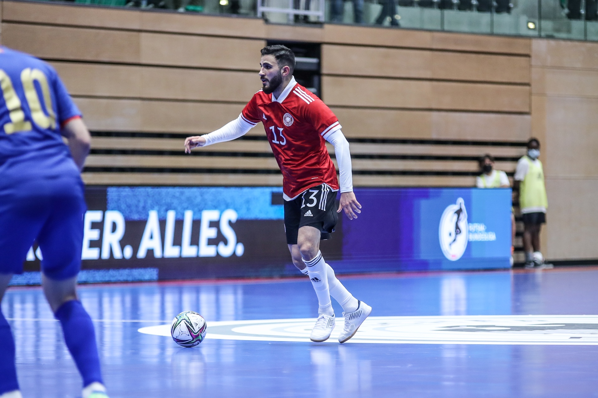 [{"clang_id":1,"value":"Futsalspieler mit Trikotnummer 13 kontrolliert Ball auf blauem Spielfeld. Im Hintergrund ein Gegenspieler mit Nummer 10 und Werbebanner."}]