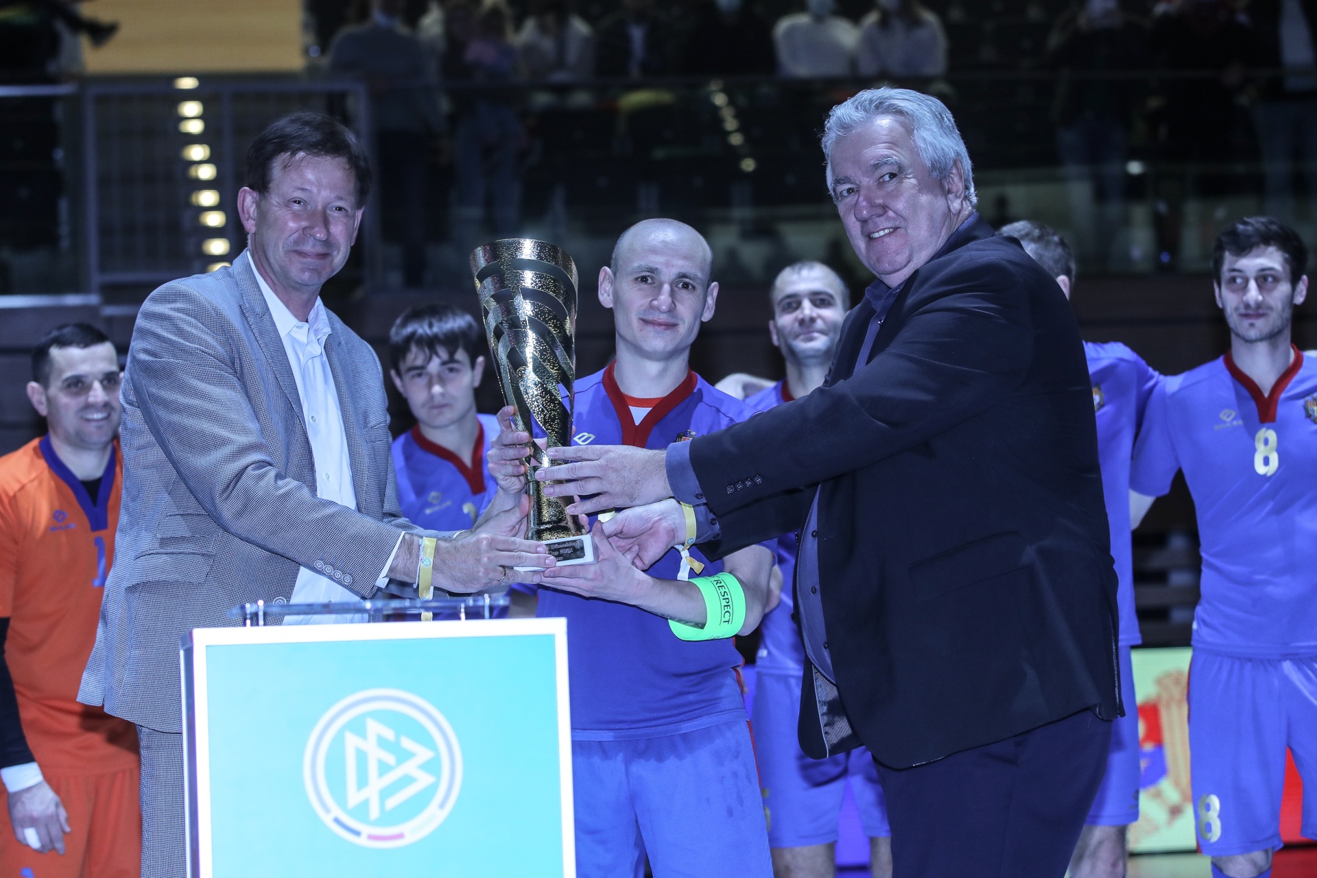 [{"clang_id":1,"value":"Zwei Männer überreichen sich eine goldene Trophäe auf einem Podest mit Logo. Im Hintergrund stehen Handballspieler in blau-roten Trikots."}]