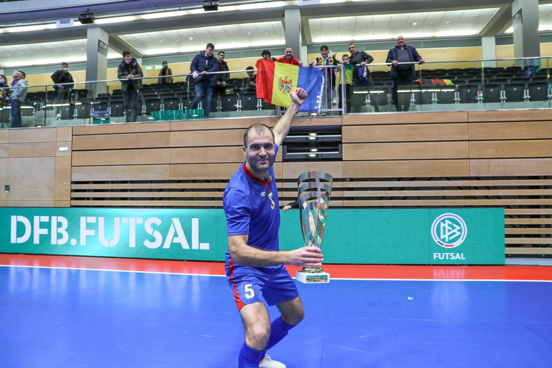 [{"clang_id":1,"value":"Futsalspieler mit Trikotnummer 5 hebt Pokal in die Höhe. Im Hintergrund Zuschauer auf einer Tribüne, Banner mit Aufschrift \"DFB.FUTSAL\". Blaue Sporthalle."}]