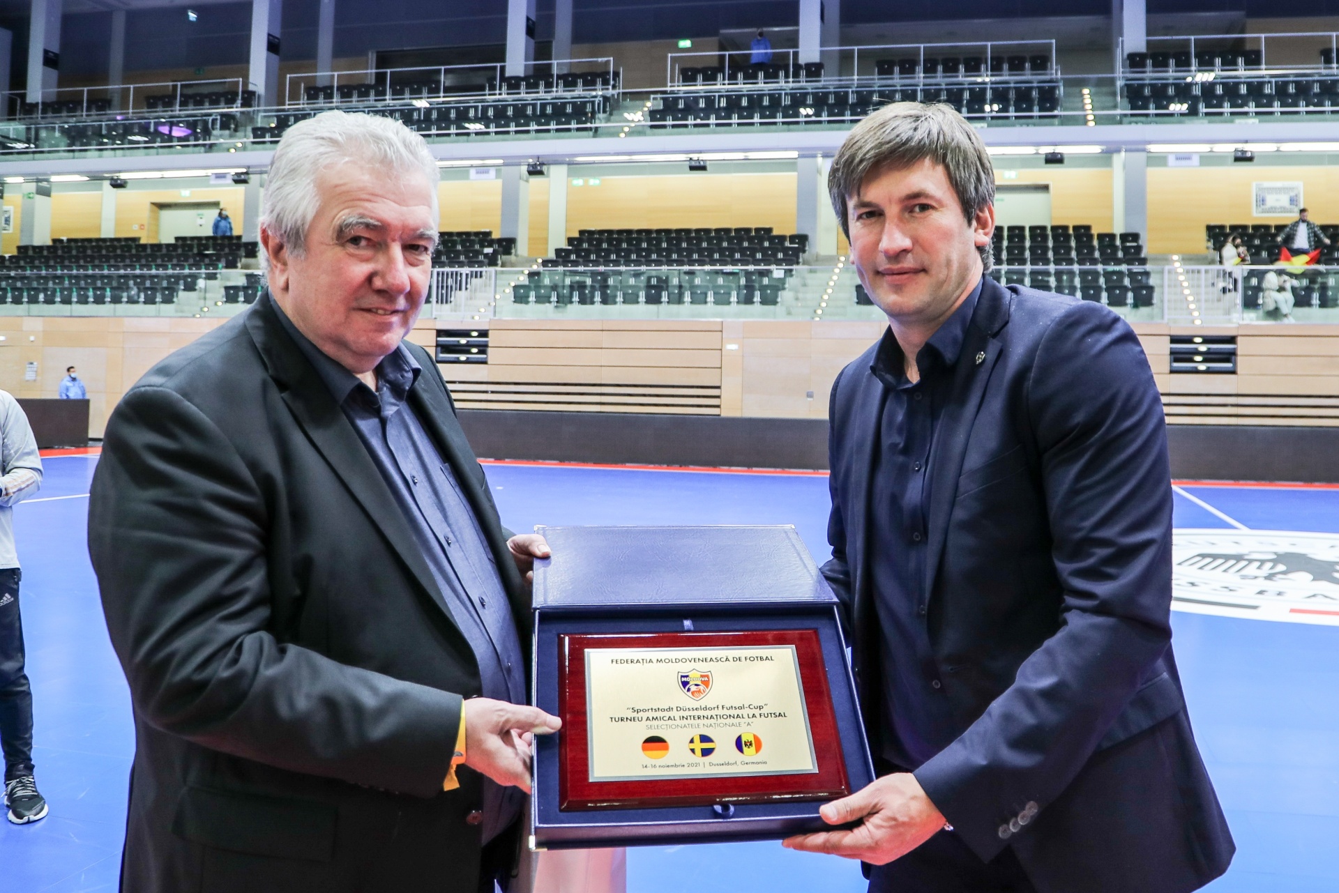 [{"clang_id":1,"value":"Zwei Männer im Anzug halten eine Auszeichnung mit Flaggen von Rumänien und Deutschland. Im Hintergrund eine leere Sporthalle mit Zuschauertribünen."}]