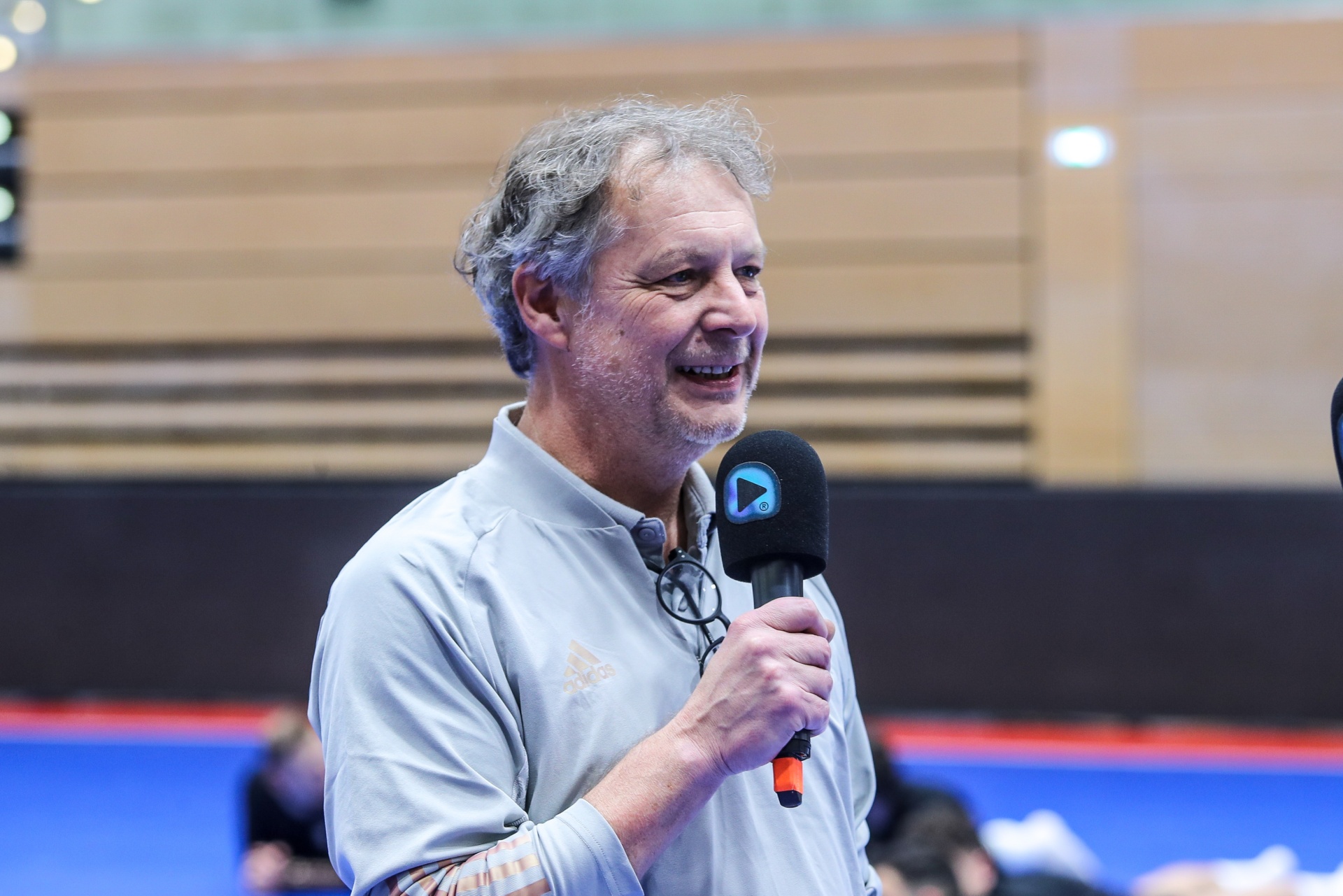[{"clang_id":1,"value":"Mann mit grauen Haaren hält Mikrofon. Er trägt ein hellgraues Sweatshirt und lächelt. Im Hintergrund eine Sporthalle mit Tribünen."}]