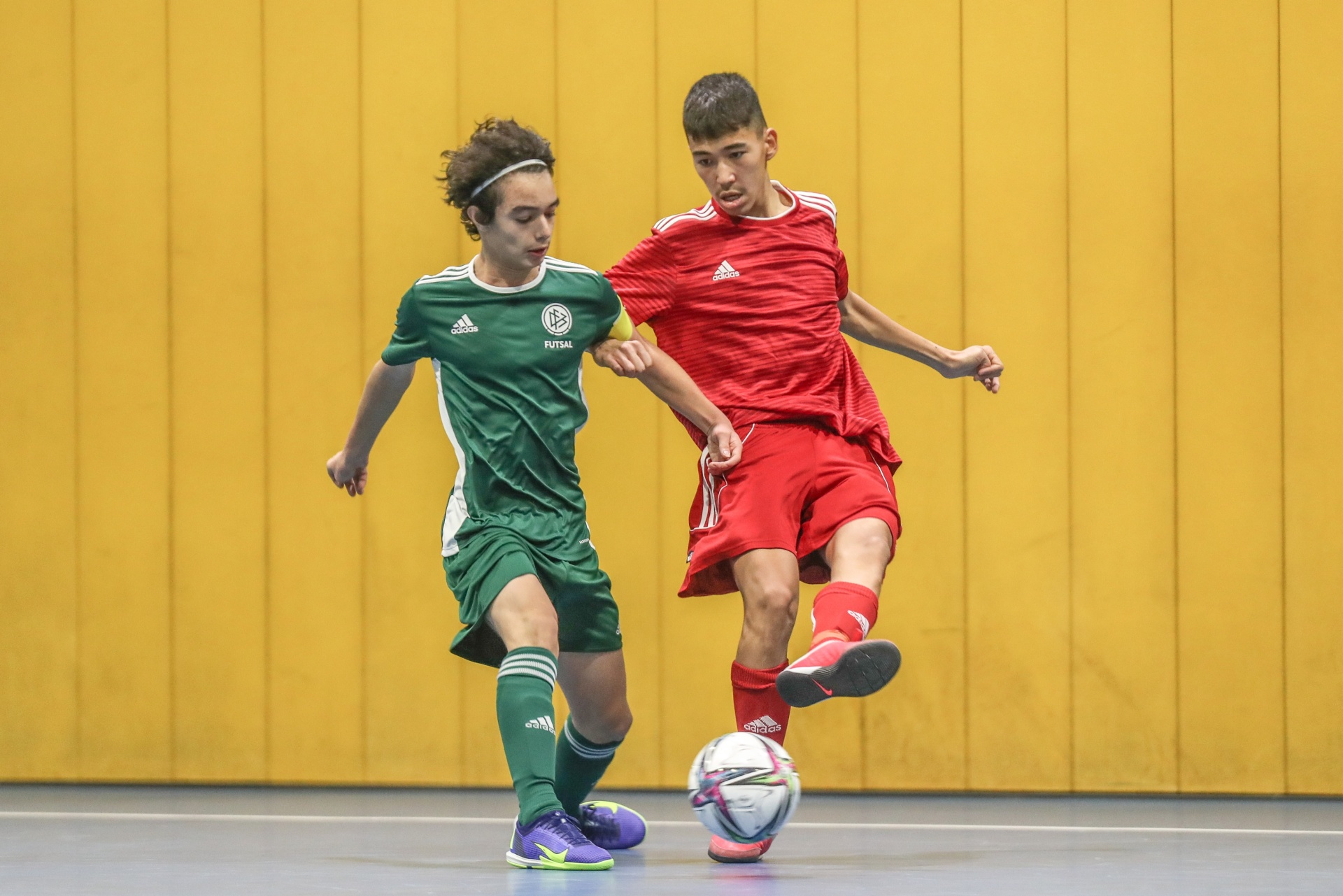 [{"clang_id":1,"value":"Zwei junge Fussballspieler im Hallensport, einer in grünem, der andere in rotem Trikot, beim Zweikampf um den Ball."}]