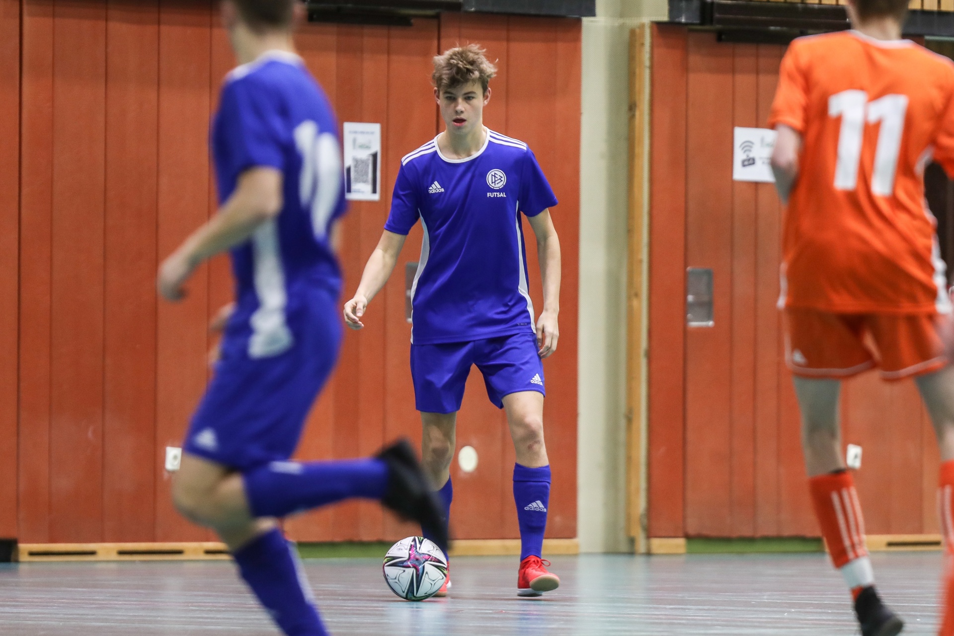[{"clang_id":1,"value":"Futsal-Spieler in blauen Trikots und Shorts sowie rote Shorts stehen auf einem Hallenfußballfeld. Ein Spieler dribbelt den Ball."}]