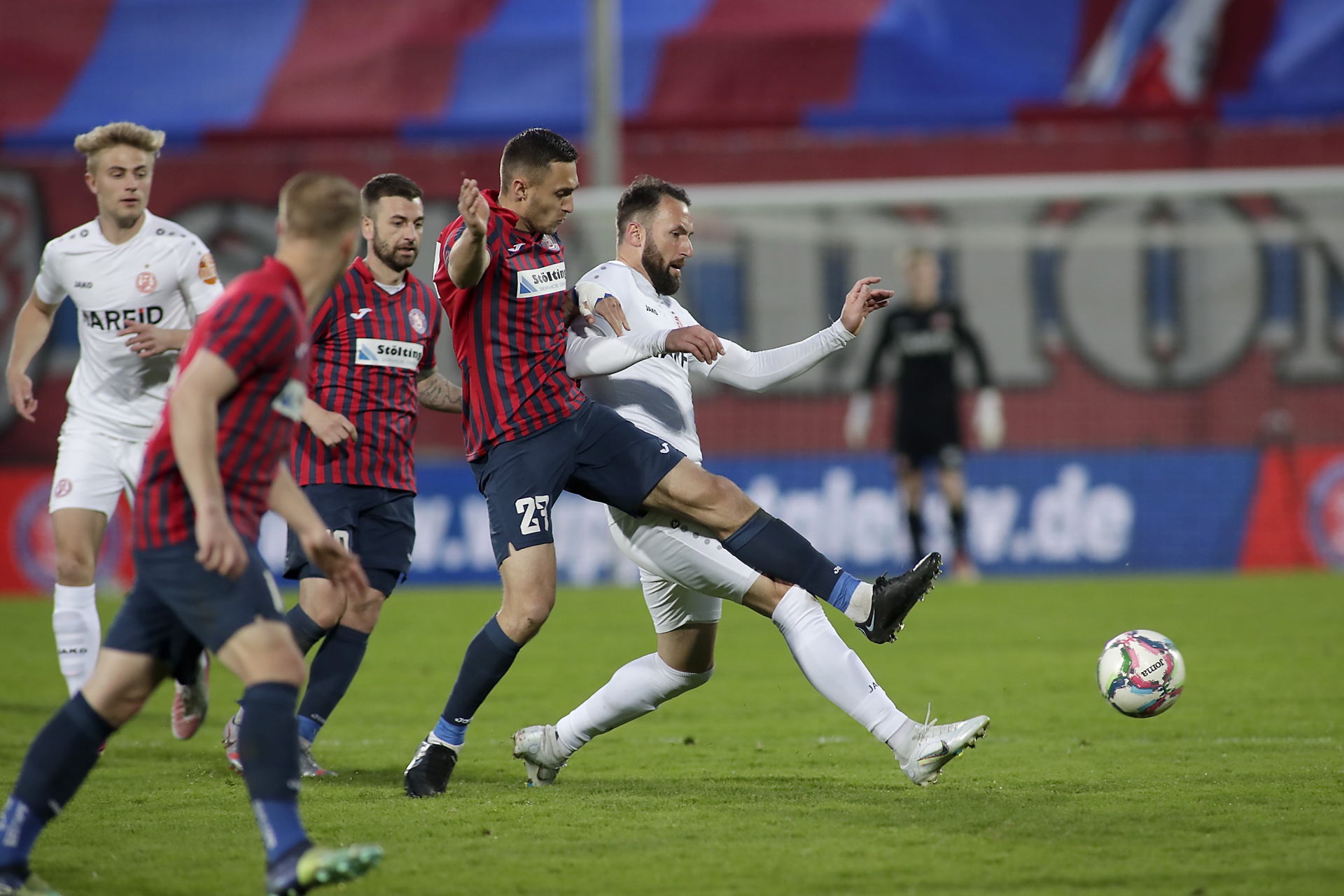 [{"clang_id":1,"value":"Fußballspiel: Zwei Spieler kämpfen um den Ball. Einer in rot-schwarzem Trikot rutscht in den Zweikampf gegen einen Spieler in weißem Trikot. Im Hintergrund weitere Spieler und Zuschauerränge."}]