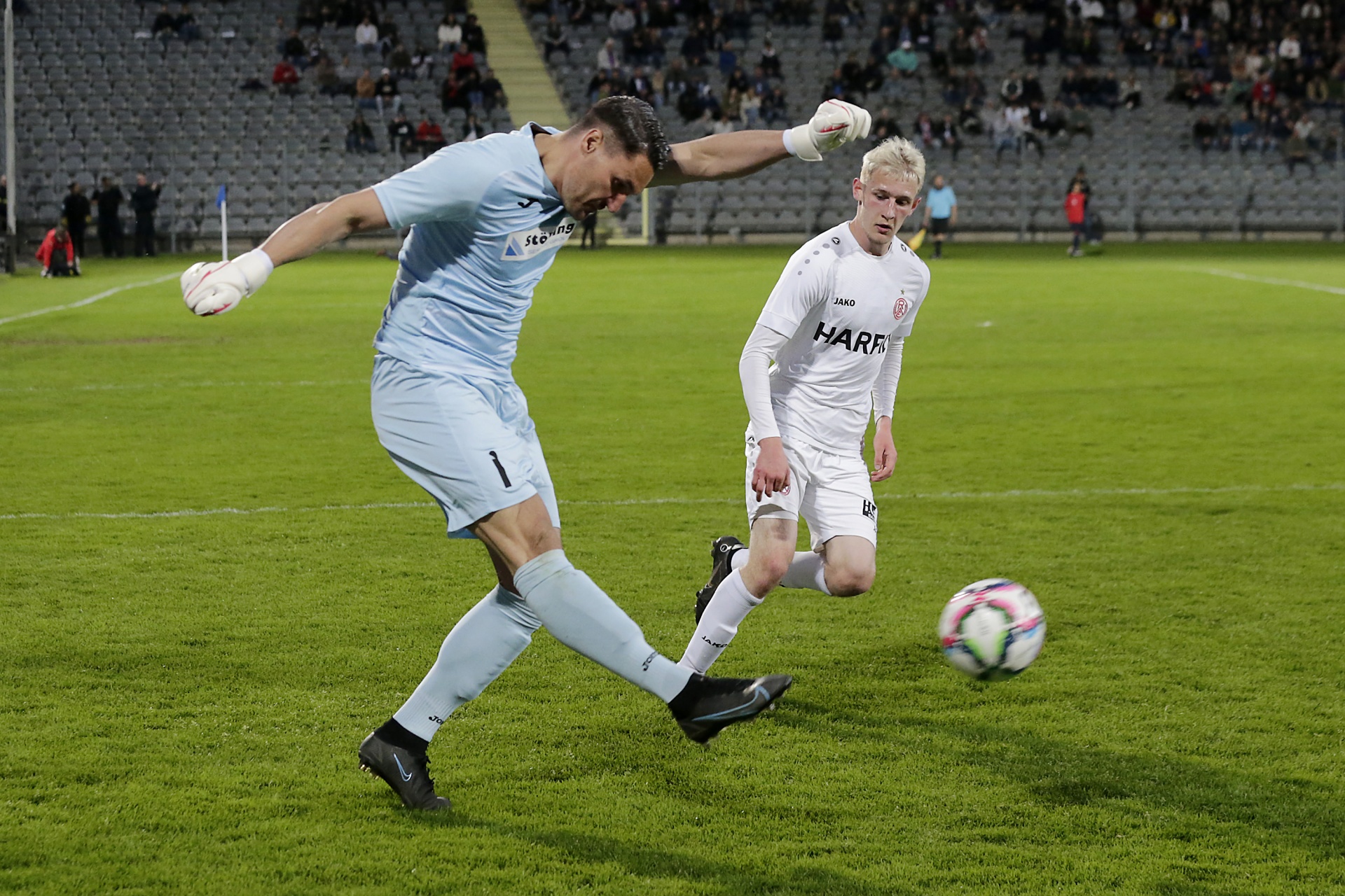 [{"clang_id":1,"value":"Fußballtorwart in blau springt zur Seite, um Schuss eines Spielers in weiß zu parieren. Rasenfeld und leere Zuschauertribünen im Hintergrund."}]