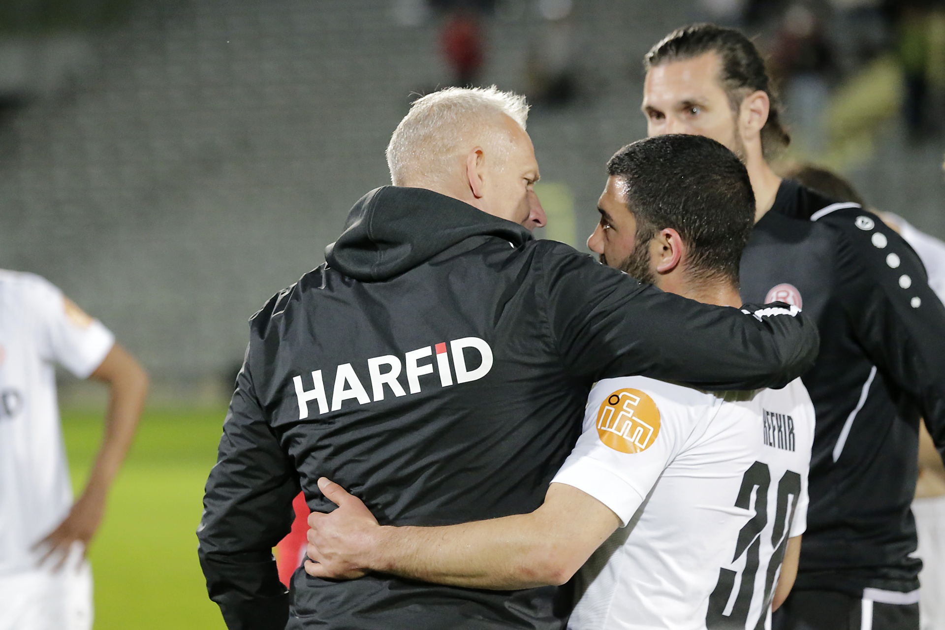 [{"clang_id":1,"value":"Zwei Männer umarmen sich auf einem Fußballfeld. Der Mann links trägt eine Jacke mit der Aufschrift \"HARFID\", der andere ein schwarz-weißes Trikot mit der Nummer 30. Im Hintergrund weitere Fußballspieler."}]