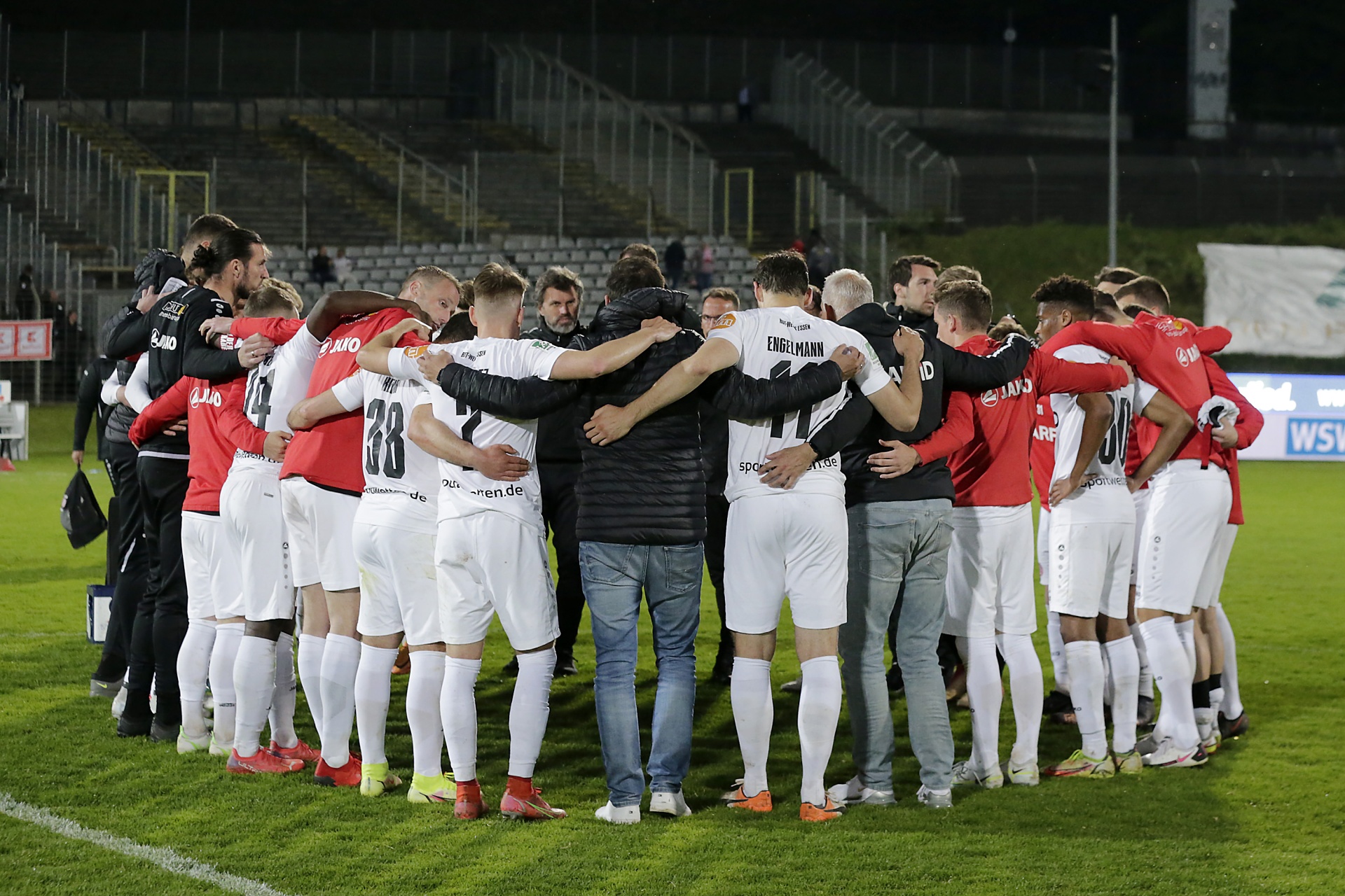 [{"clang_id":1,"value":"Fußballmannschaft im Kreis auf dem Spielfeld. Spieler tragen rote und weiße Trikots, kurze Hosen und Stutzen. Im Hintergrund leere Zuschauertribüne."}]