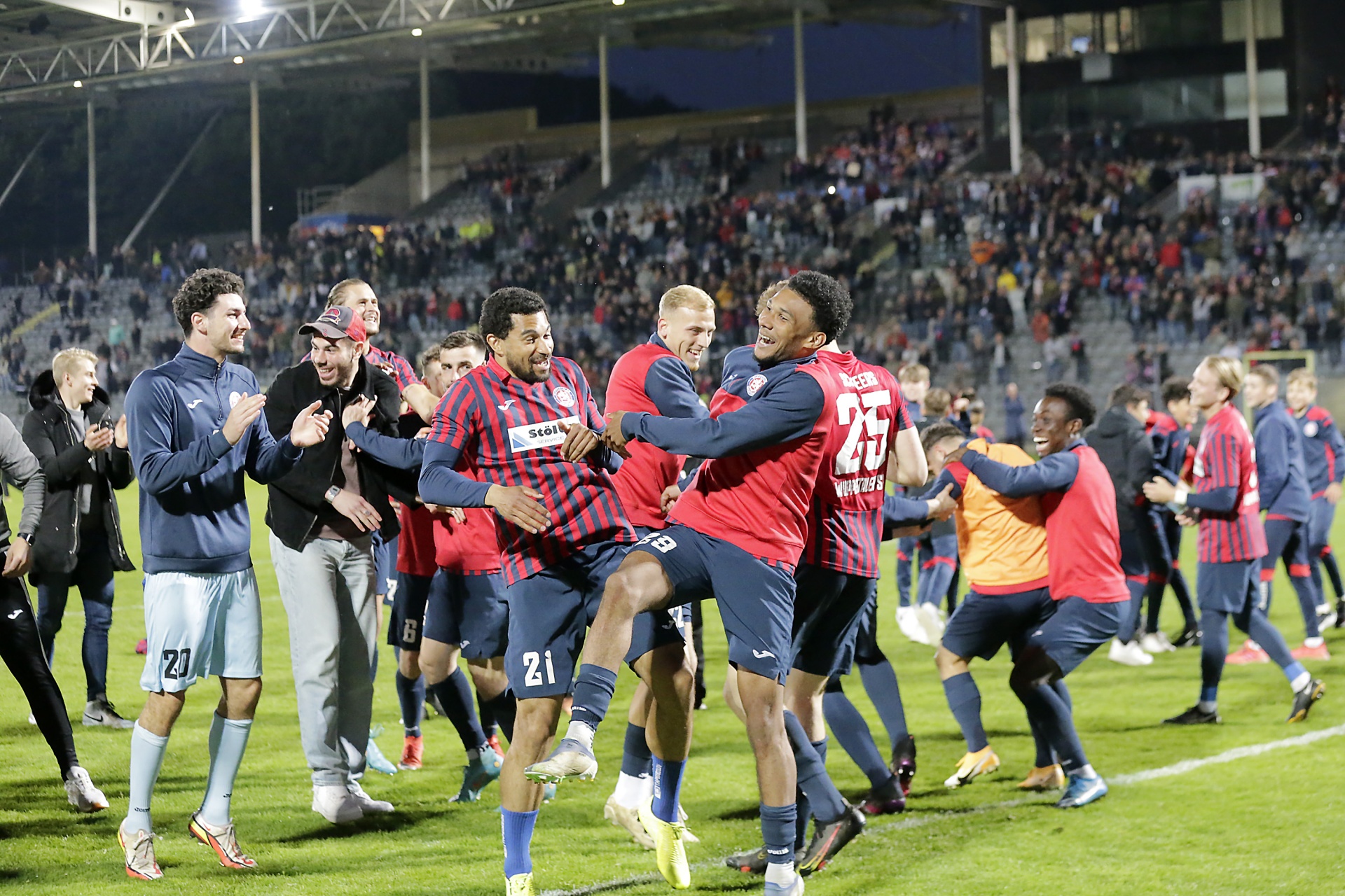 [{"clang_id":1,"value":"Fußballspieler in roten und blauen Trikots feiern auf dem Spielfeld, umringt von Zuschauern im Stadion. Einige Spieler werden von Teamkollegen gehoben."}]