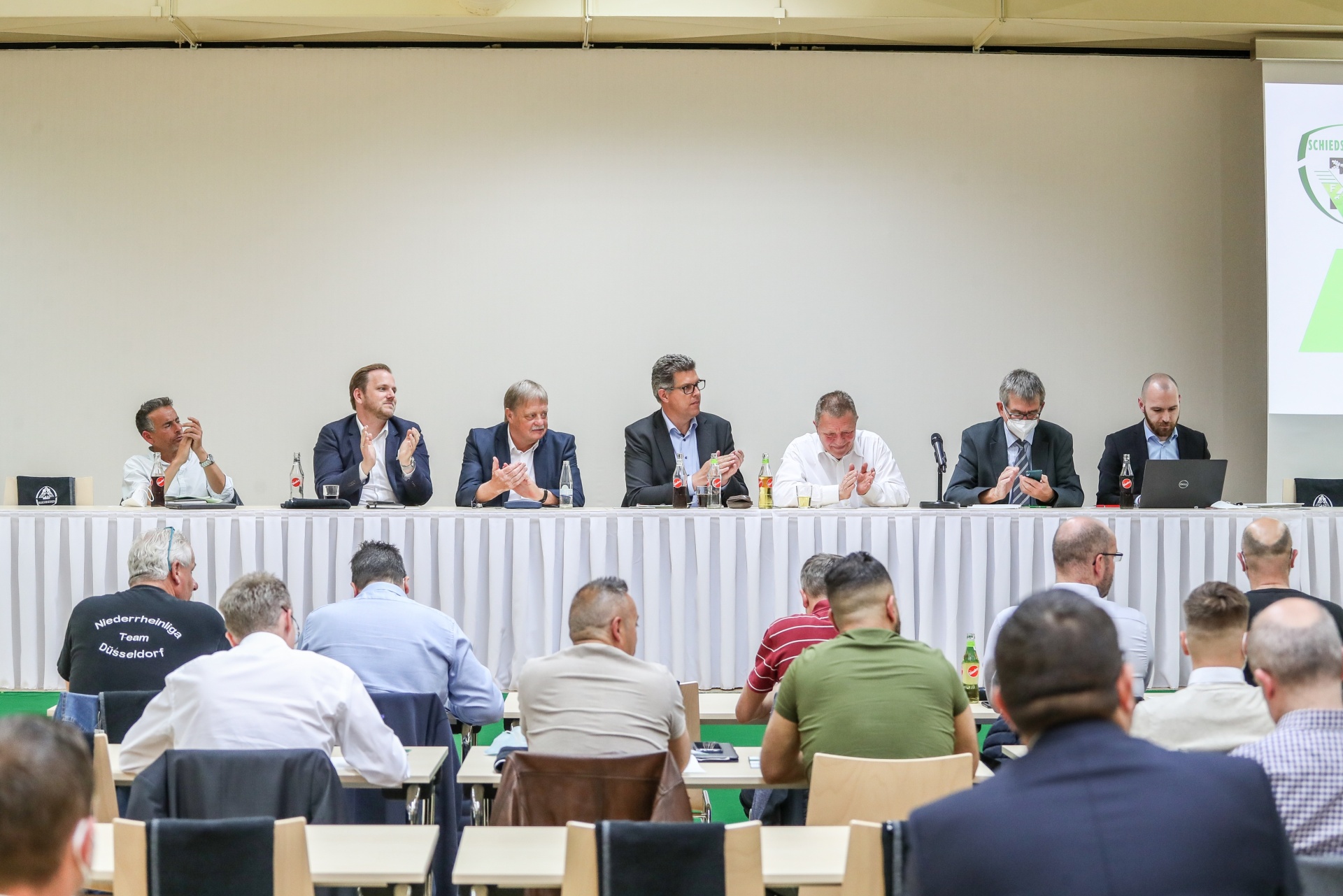 [{"clang_id":1,"value":"Konferenz: Am Tisch sitzen sieben Männer, diskutieren und schauen in die Mitte. Im Publikum sind weitere Personen zu sehen."}]