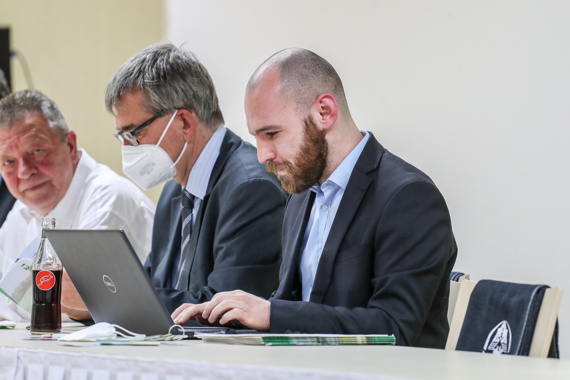 [{"clang_id":1,"value":"Drei Männer sitzen an einem Tisch. Der Mann im Vordergrund arbeitet auf einem Laptop, trägt Anzug und Krawatte. Neben ihm sitzt ein Mann mit Maske. Eine Cola-Flasche steht auf dem Tisch."}]