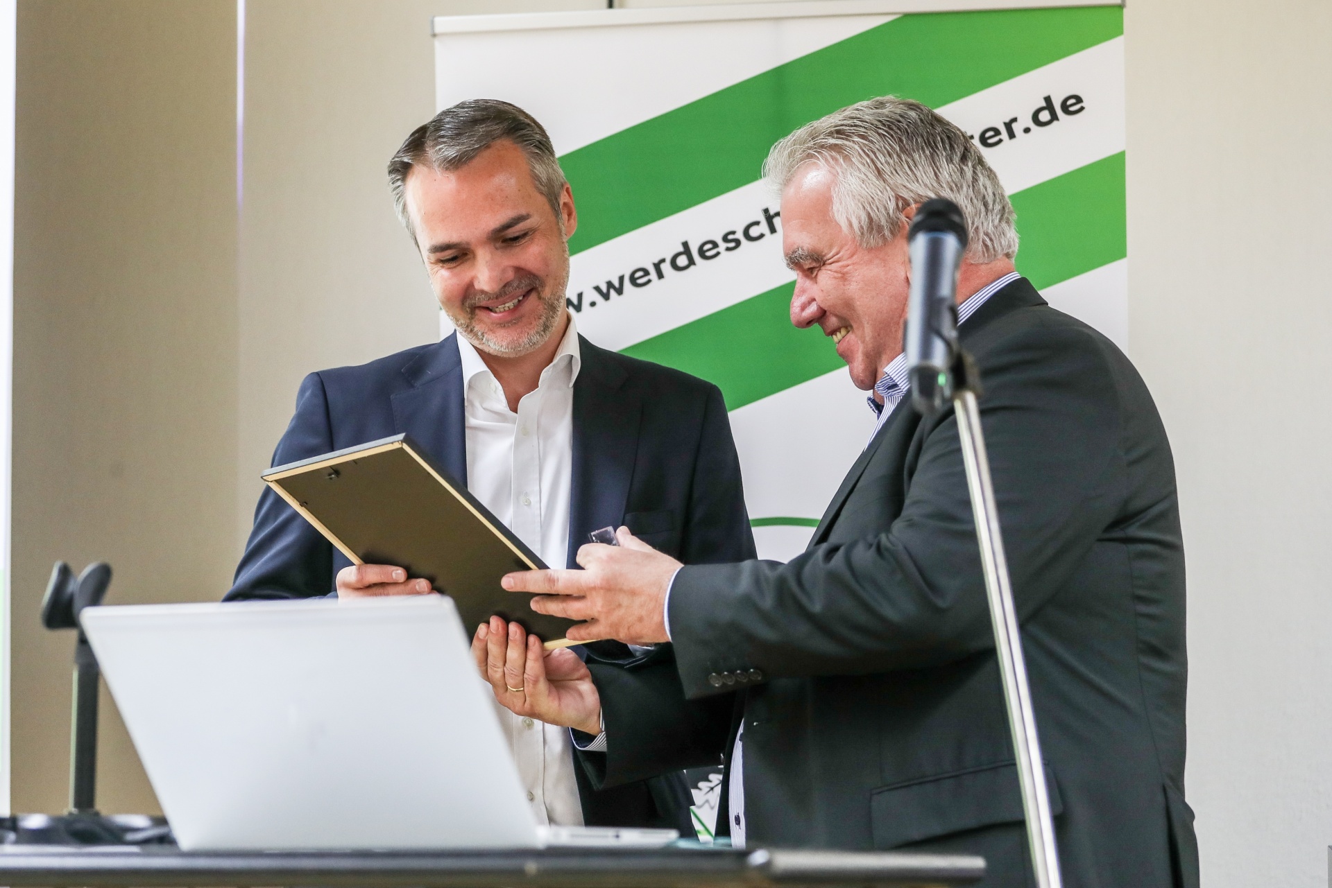 [{"clang_id":1,"value":"Zwei Männer in Anzug mit Dokument, einer übergibt es dem anderen. Im Hintergrund ein Banner mit grün-weißer Grafik und Schriftzug."}]