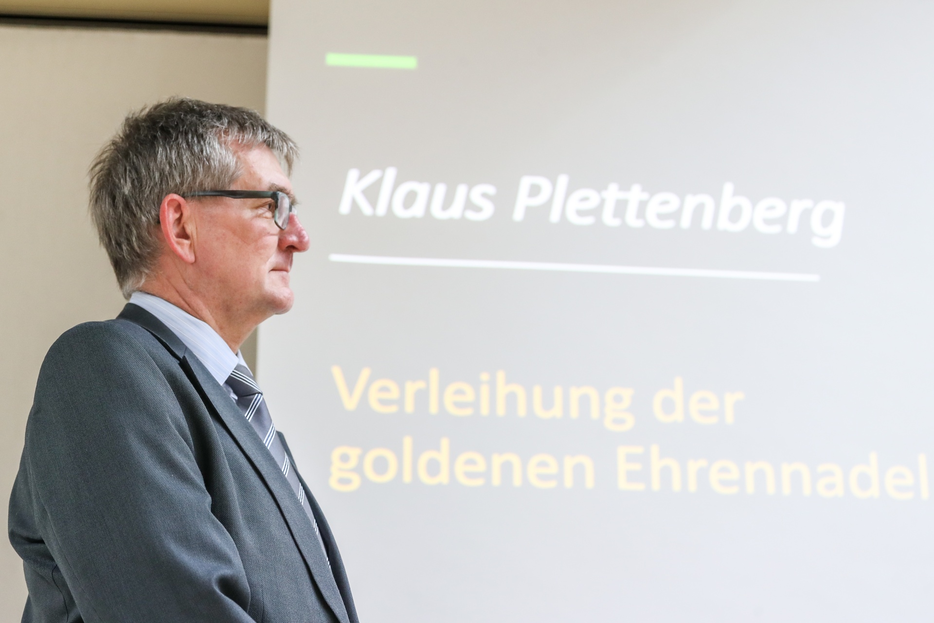 [{"clang_id":1,"value":"Mann mit Anzug und Brille steht seitlich vor einer Projektionsfläche. Darauf steht \"Klaus Plettenberg\" und \"Verleihung der goldenen Ehrennadel\"."}]