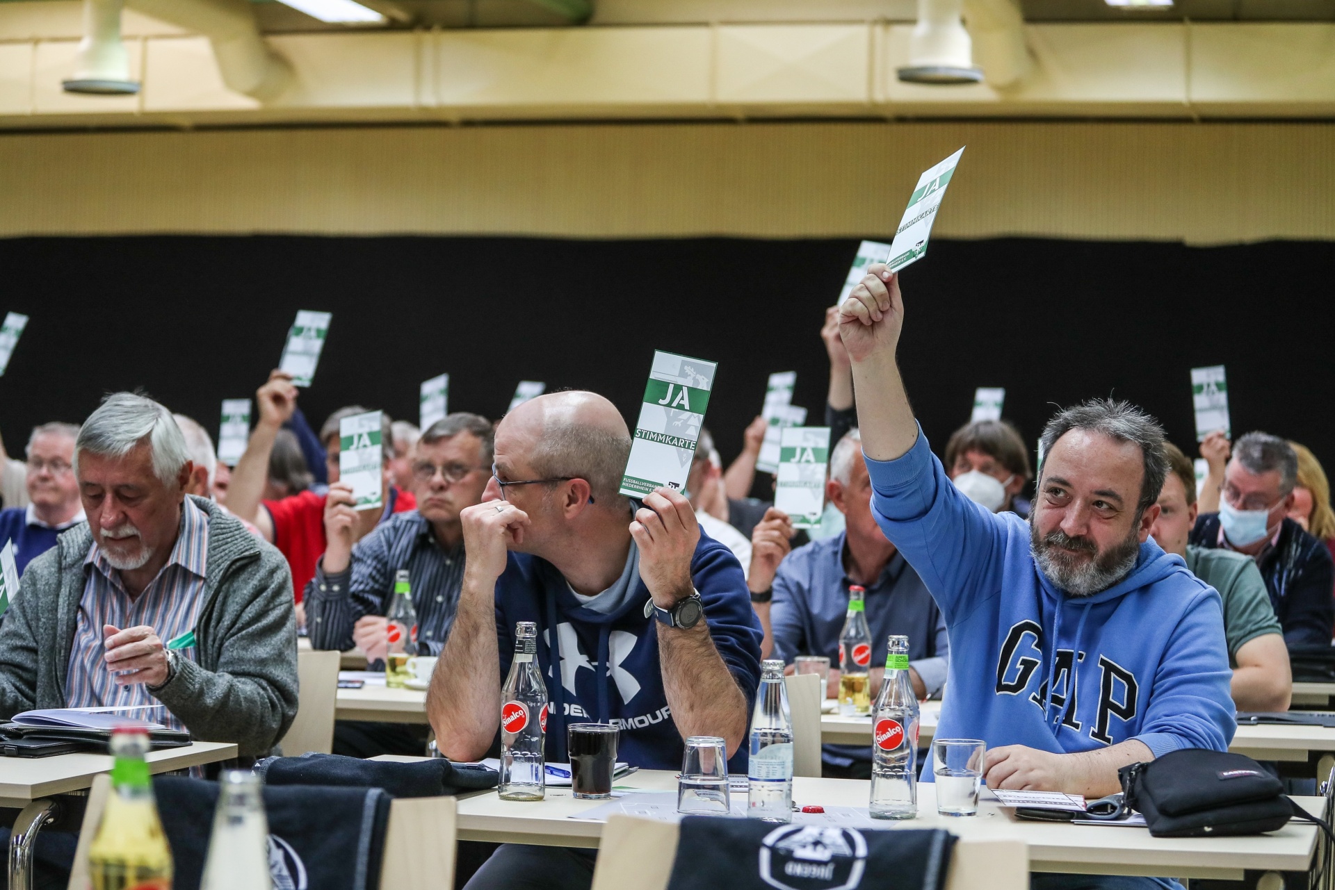 [{"clang_id":1,"value":"Versammlung von Männern an Tischen. Einige heben grüne Karten hoch, andere schreiben oder blicken nach vorn. Getränkeflaschen und Notizblöcke auf den Tischen."}]